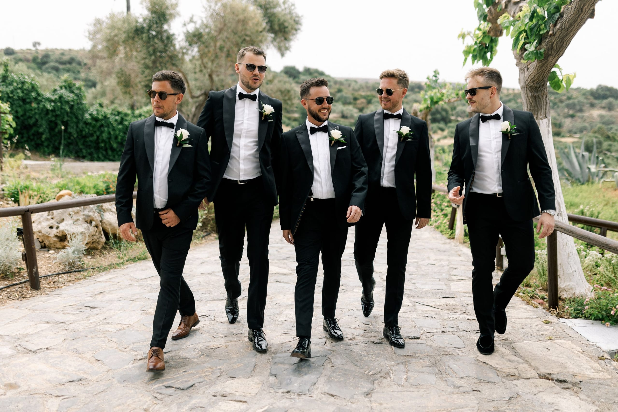 Five men in black tuxedos with bow ties and white boutonnieres of white roses walk together along a flagstone path at AgrecoFarms in Adele, Crete, Greece, past a wooden rail and olive trees. Most wear dark sunglasses, and the man in the centre is smiling broadly as he strides forward, while the others look ahead or to the side in relaxed conversation. The warm, soft daylight and green hills behind them give the wedding-day scene a calm, convivial mood.