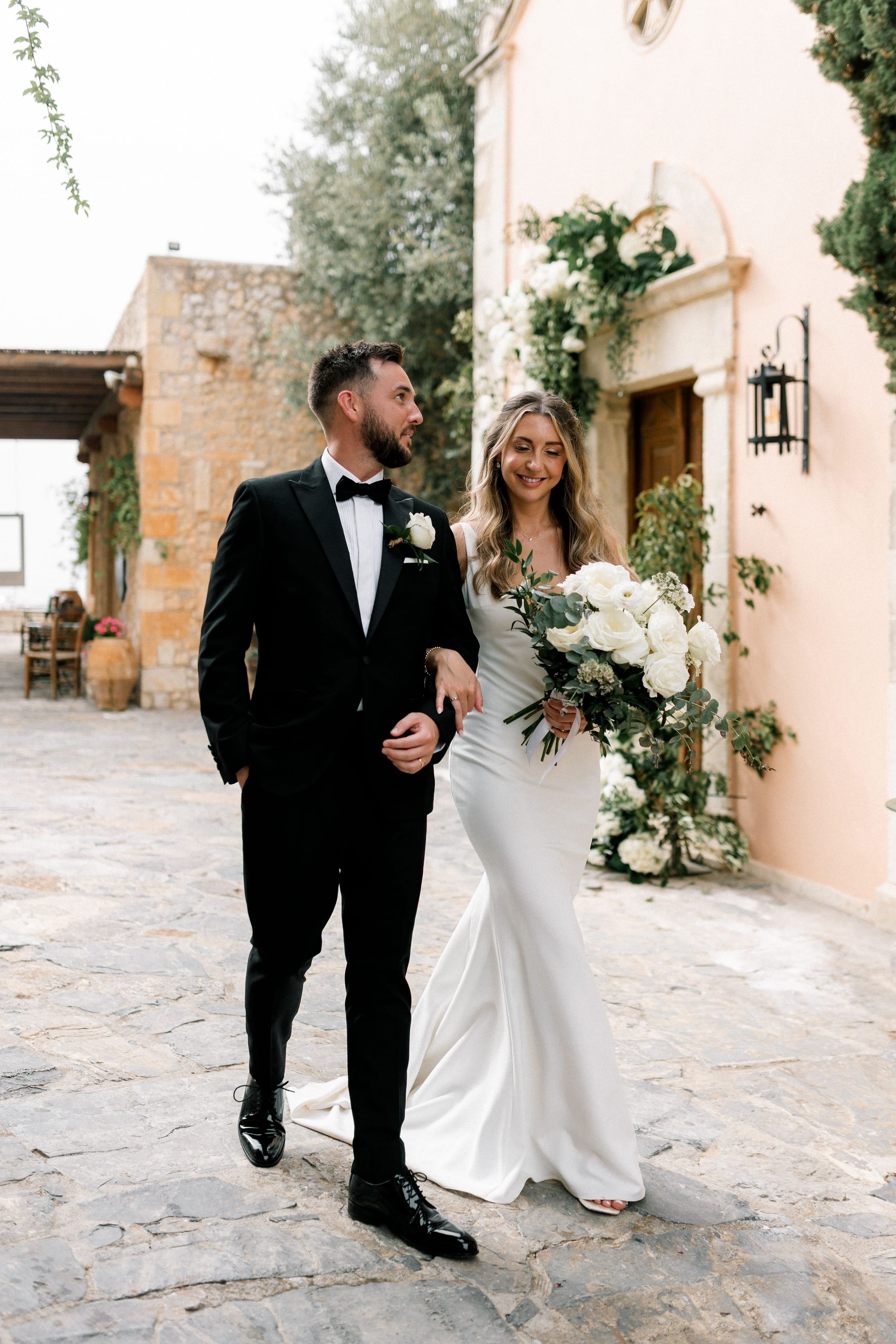A bride in a fitted white satin gown and a groom in a black tuxedo with a white rose boutonniere stroll arm in arm along a cobbled courtyard at AgrecoFarms in Adele, Crete, Greece. She carries a loose bridal bouquet of white garden roses and eucalyptus, and a white rose floral arrangement frames the pale pink wall and wooden door behind them. Soft, natural light and olive trees and stone walls create a calm, romantic mood.