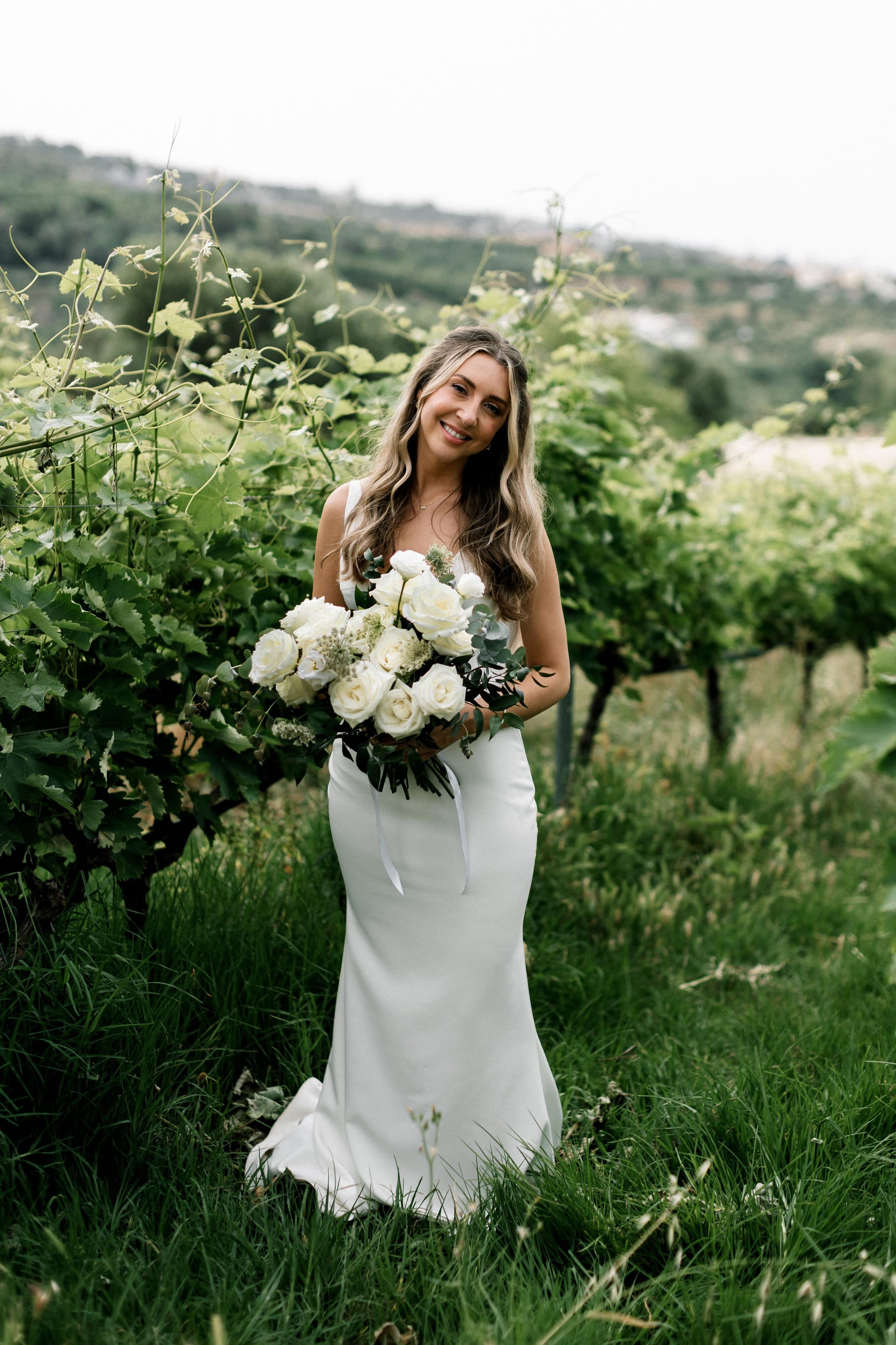 A bride in a sleek white satin gown stands smiling among leafy grapevines and tall green grass, holding a full, rounded bouquet of white garden roses trimmed with eucalyptus and delicate white filler blooms. The soft, overcast light and hazy hills behind her give the scene a calm, airy mood. This is a wedding bouquet photographed at AgrecoFarms in Adele, Crete, Greece.