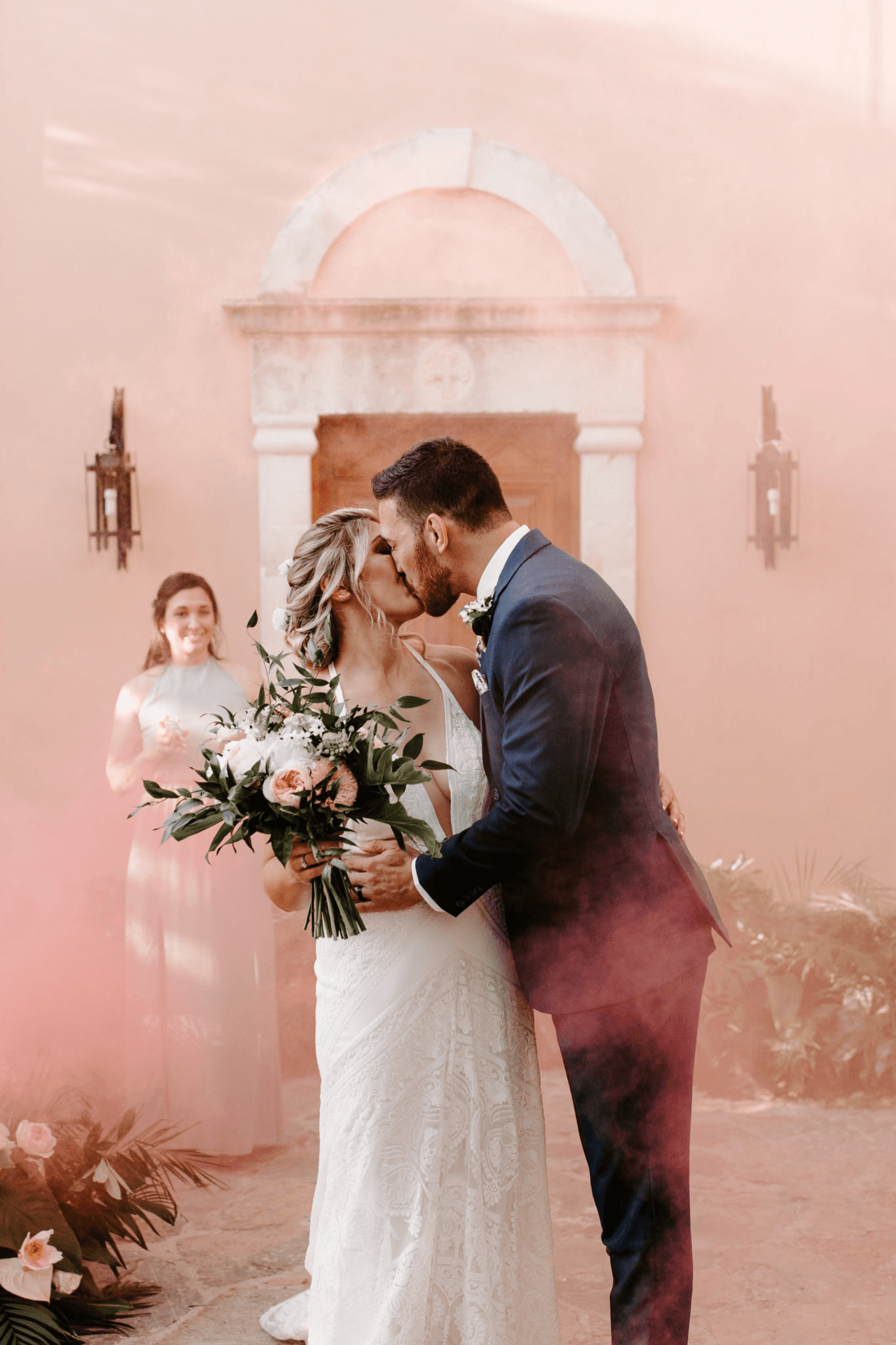 A bride and groom share a kiss in a wash of pink smoke in front of a rose-coloured wall with a wooden door and lanterns, at a wedding in Greece. The bride, in a lace gown with a deep V neckline, holds a bouquet of blush garden roses, white blooms and trailing green eucalyptus. The groom wears a navy suit with a white boutonniere, and a smiling bridesmaid in a pale blue dress watches from the left.