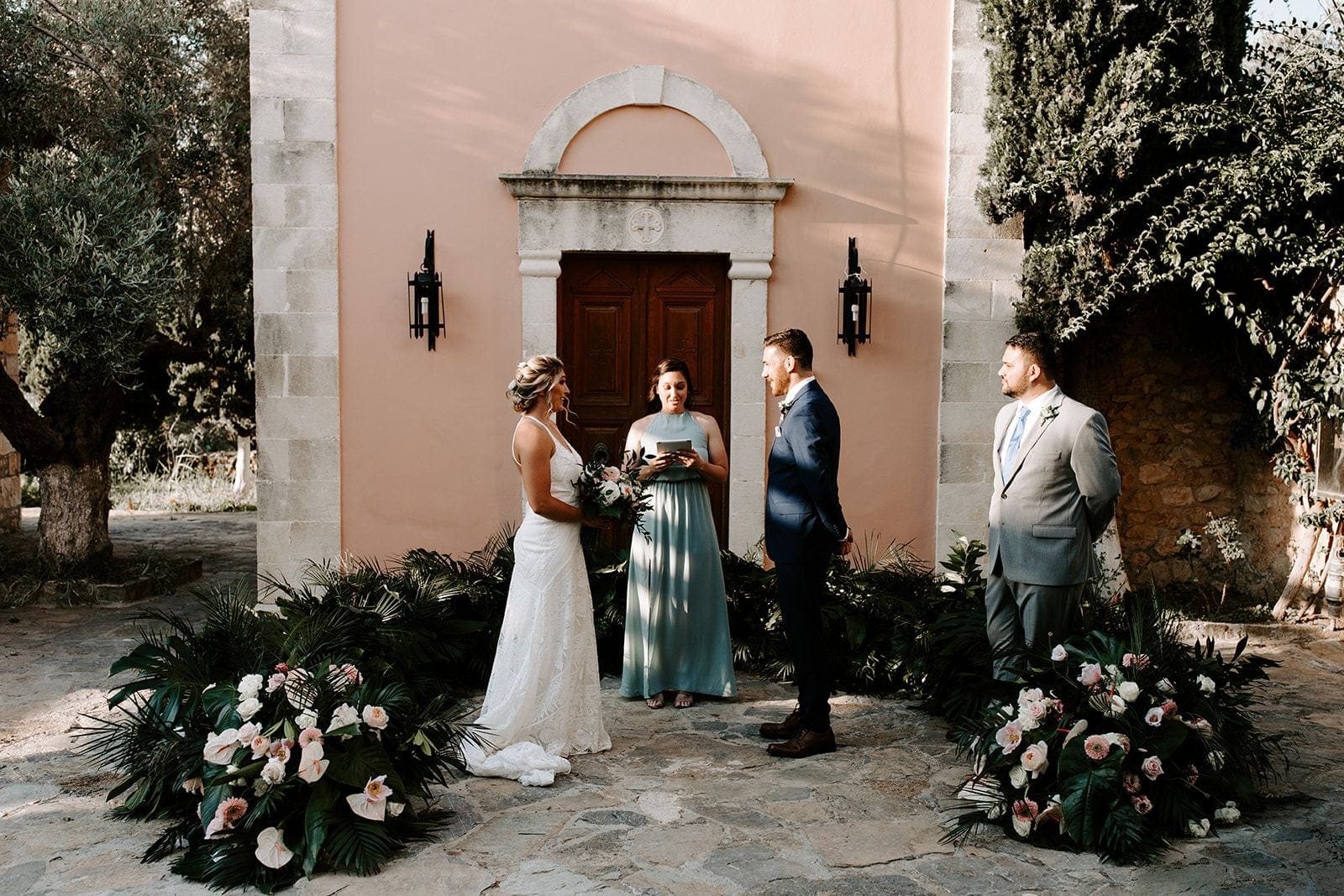 Two palms-and-blush floral installations frame a small wedding ceremony in front of a peach-washed chapel with a carved stone doorway and a dark wooden door, in Greece. The bride in a lace gown holds a loose bouquet of pale pink and white blooms, while the groom in a navy suit faces her and a celebrant in a dusty-blue gown reads from a tablet. Warm late-afternoon light falls softly across the stone courtyard.