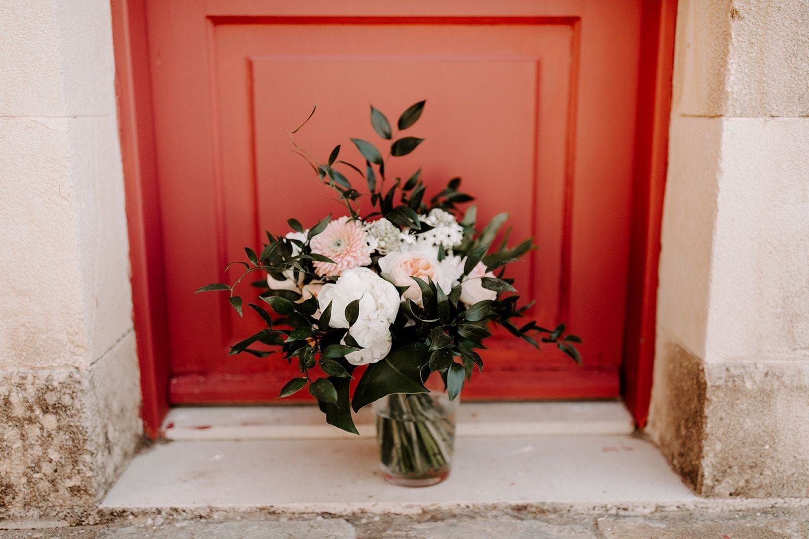A bridal bouquet of white peonies, blush garden roses, a pale pink dahlia and tiny white blooms, tied with trailing dark green foliage, sits in a clear glass vase on a pale stone step in front of a rust-red wooden door. The soft, natural light and muted palette give it a calm, romantic feel, a wedding flower arrangement photographed in Greece.