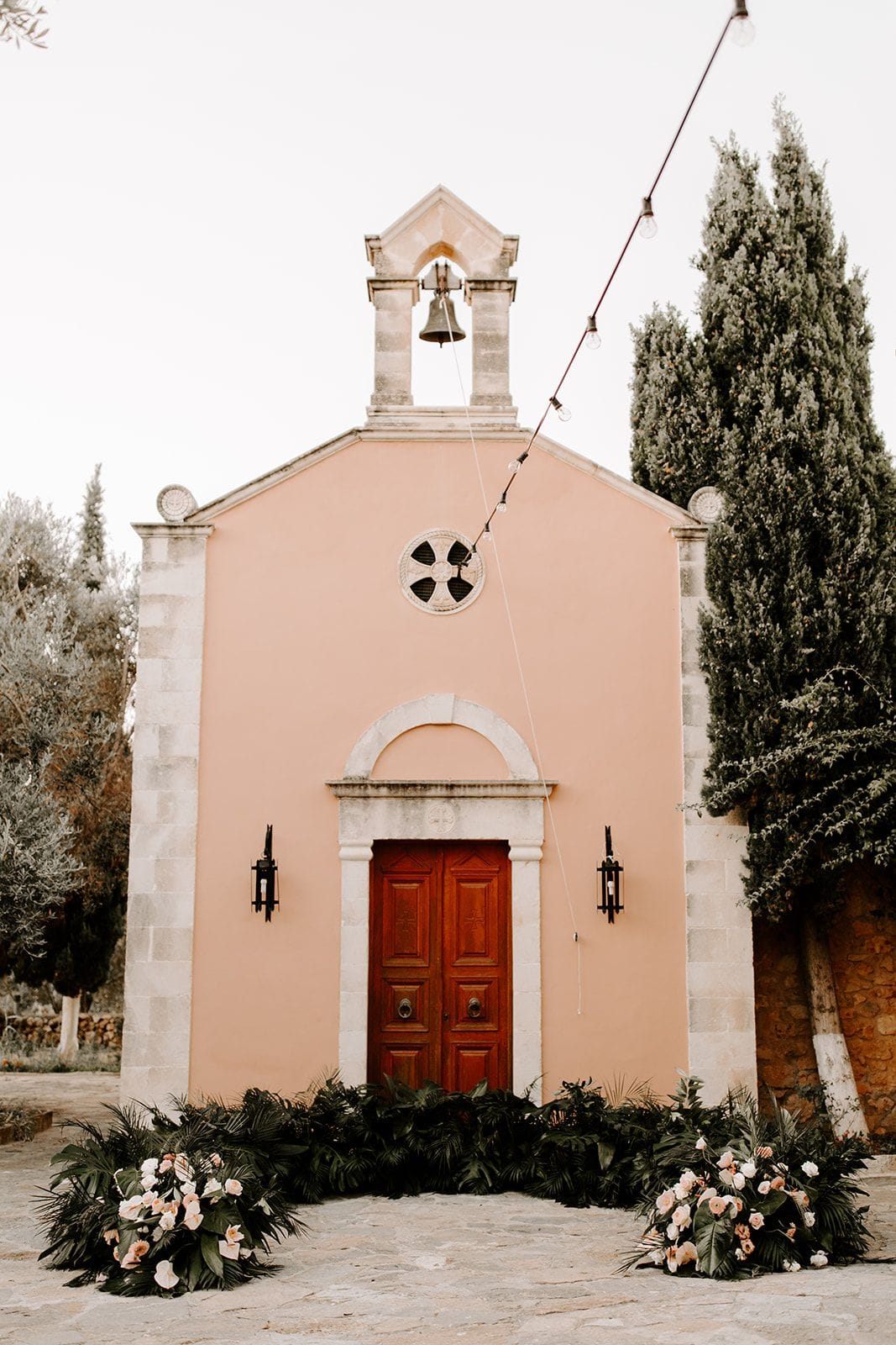 Two low floral arrangements of pale pink and peach roses, calla lilies and blush blooms sit on stone paving at either side of a dark green bed of palm and monstera foliage, framing the entrance of a peach-washed church in Greece. Behind them, a pair of arched wooden doors is flanked by black iron lanterns, and tall cypress trees and olive branches soften the soft, warm light. The scene is a wedding ceremony setting in Greece, with the flowers arranged as a welcoming path to the church door.