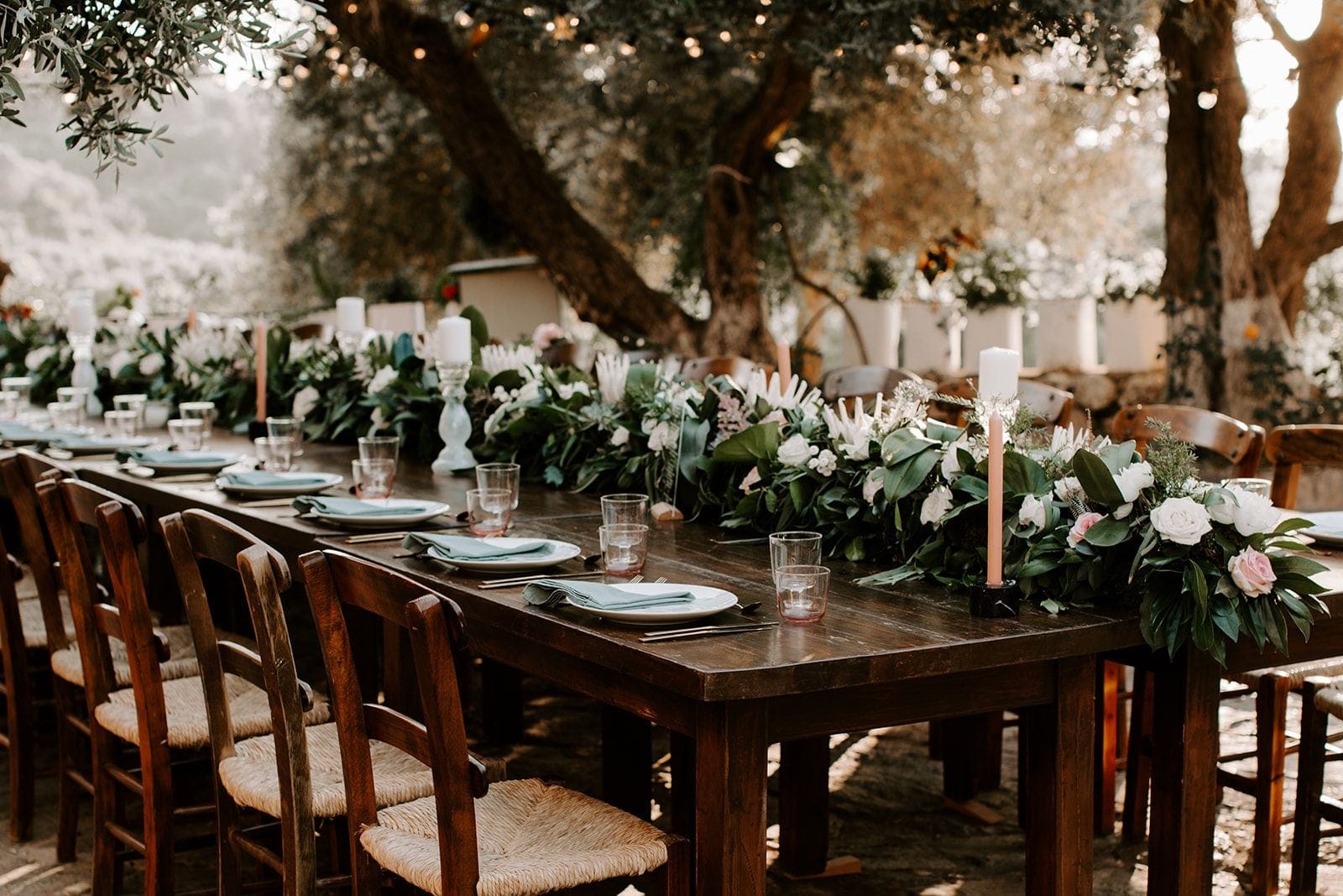 A long rustic wooden table under old olive trees, dressed with a lush garland of white garden roses, blush pink roses, white protea and trailing greenery, with tall blush taper candles rising from the centre. Place settings with white plates and sage green napkins sit beneath strings of warm lights, creating a soft, golden wedding table scene in Greece.