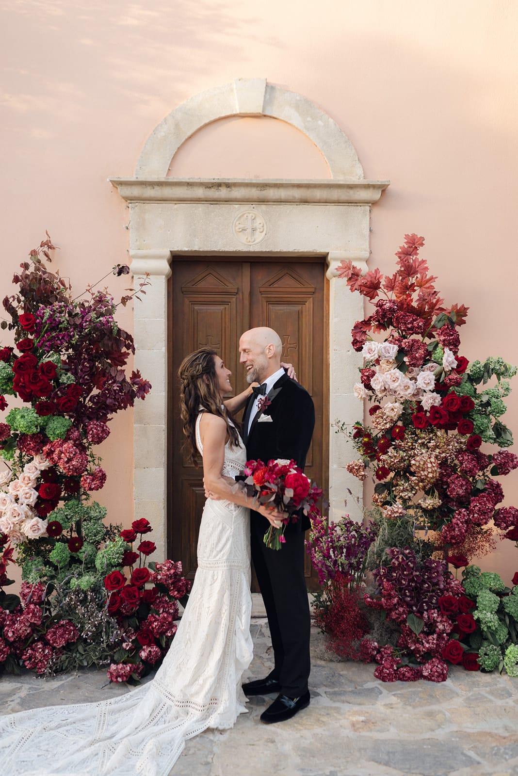 A bride in a lace-paneled ivory gown and a groom in a black velvet tuxedo embrace and smile at each other in front of a wooden double door, framed by towering floral arches of deep red roses, burgundy hydrangeas, cream roses and green hydrangeas. The bride holds a bouquet of red peonies and dark foliage. This is a wedding flower display at Agreco Farm in Greece, with a warm, romantic mood.