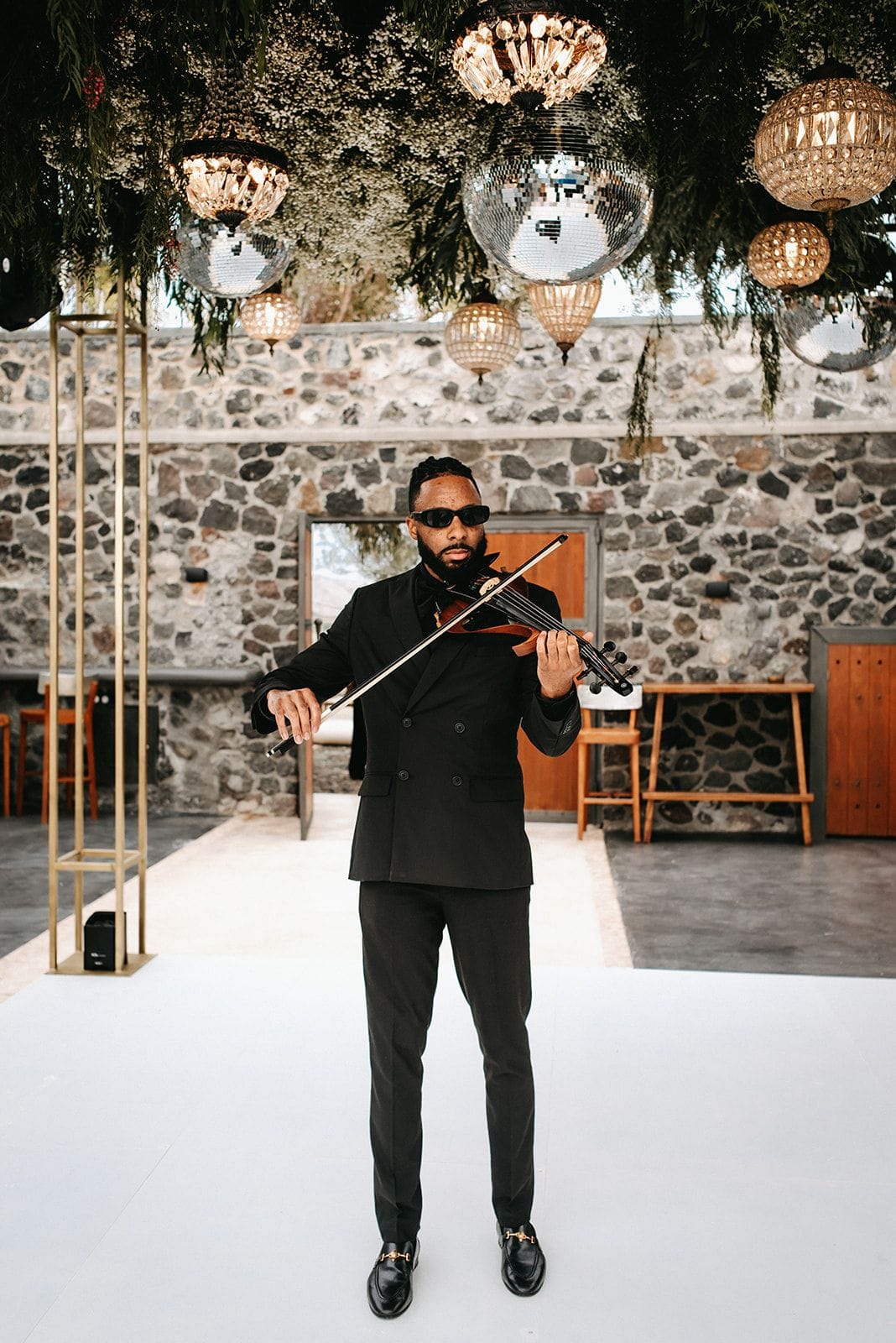 A violinist in a black double-breasted suit and dark sunglasses stands on a white floor, playing beneath a canopy of trailing greenery and small white blossoms strung with crystal chandeliers and mirror balls, part of a wedding flower installation at Cavo Ventus Luxury Villa in Santorini, Greece. Behind him, a rough stone wall and wooden doors give the room a warm, rustic character.