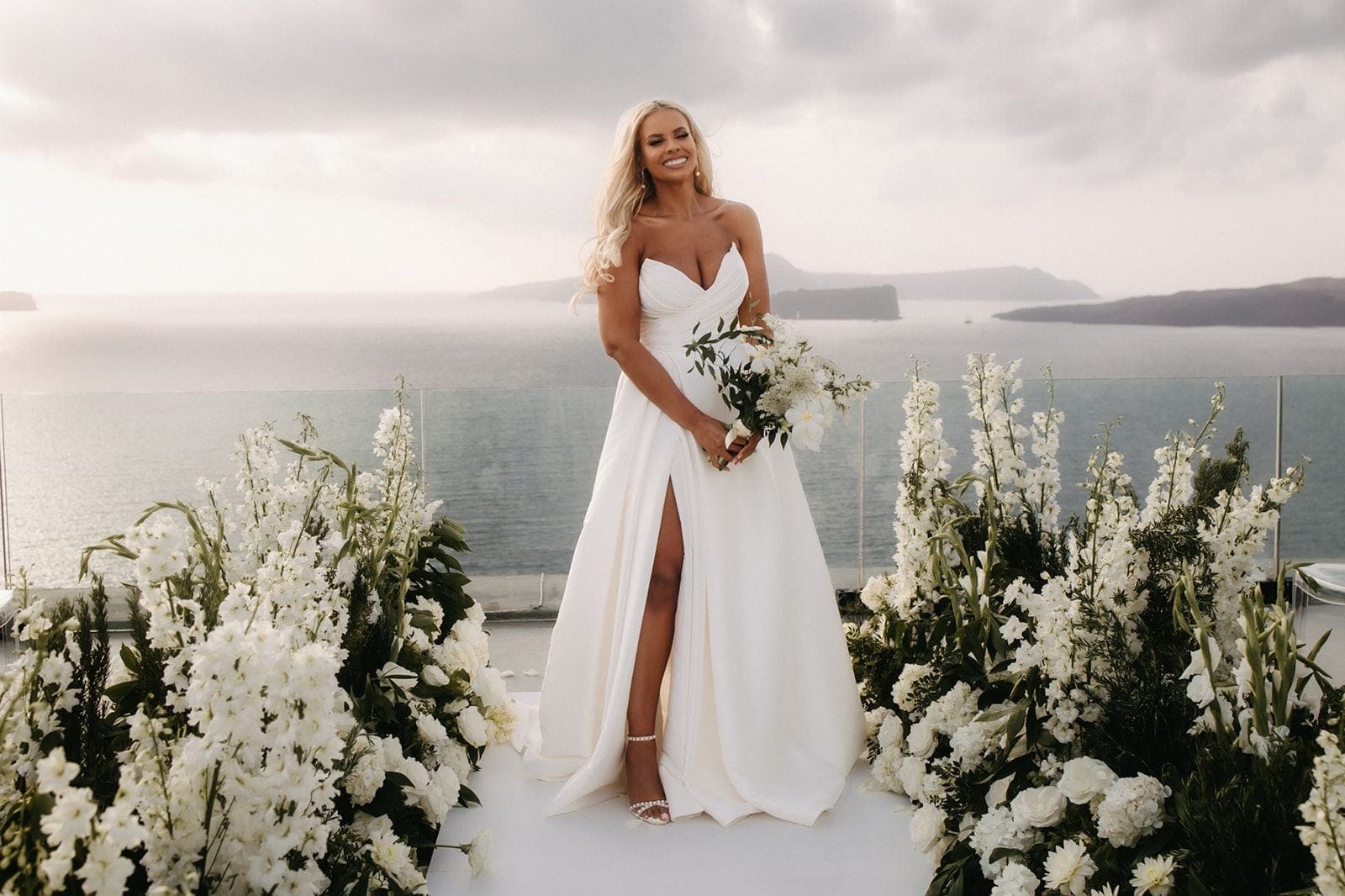 A bride in a strapless white gown with a high thigh slit stands smiling on a white aisle at Cavo Ventus Luxury Villa in Santorini, Greece, holding a loose bouquet of white blooms and greenery. On either side, tall white delphinium spires and full white garden roses frame the path, with dark green foliage tucked between them. Behind her, a glass railing gives way to the calm sea and hazy islands under a soft, overcast sky.