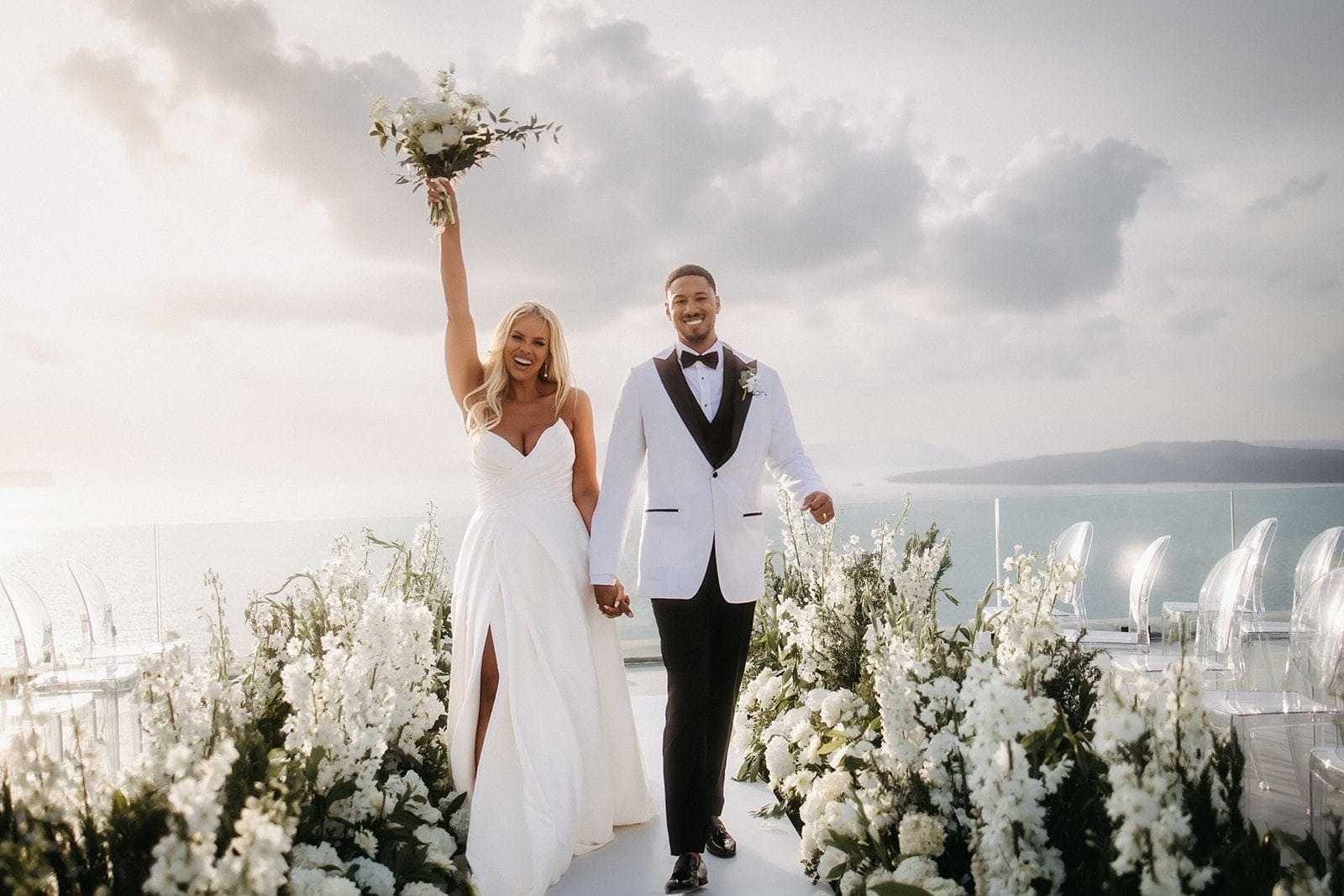 A bride in a white satin gown with a thigh-high slit raises a white bouquet of roses and eucalyptus high in one hand while holding hands with a groom in a white dinner jacket and black bow tie. They walk along a white aisle lined with tall white delphinium and hydrangea, with clear acrylic chairs beyond and the sea and distant hills under a soft cloudy sky, at Cavo Ventous, a wedding venue in Greece.
