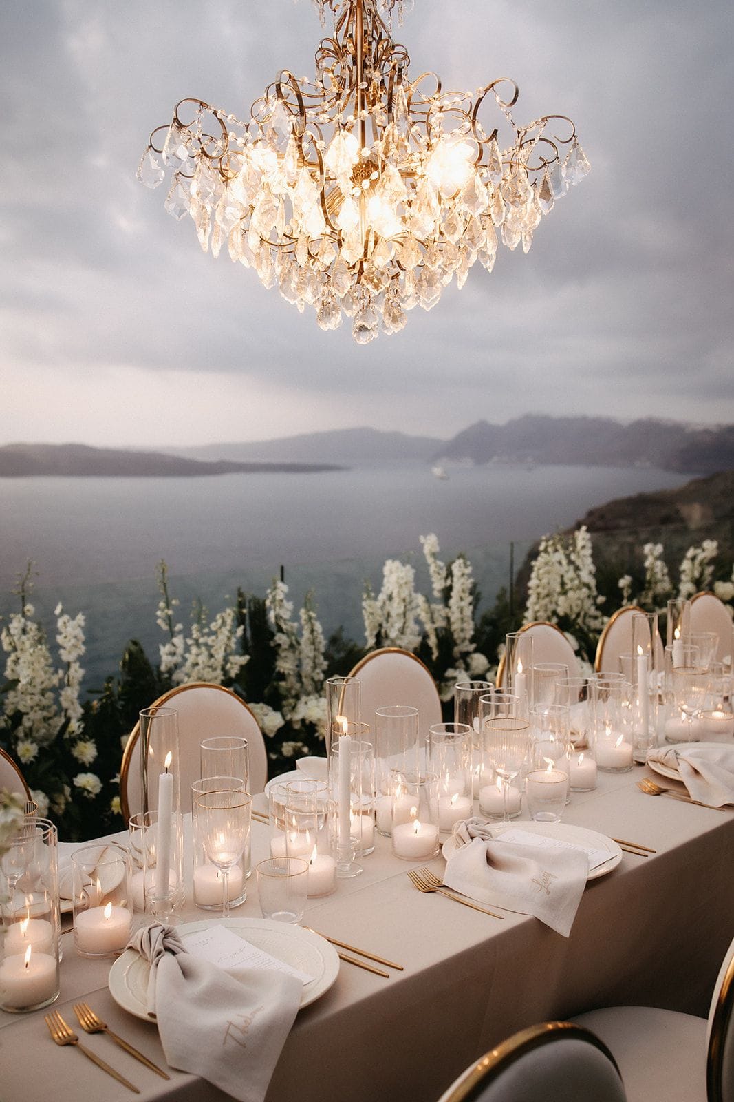 A long wedding dinner table dressed in cream linen, with gold-rimmed plates, gold cutlery and linen napkins, lit by rows of pillar and tealight candles in glass hurricanes, all set beneath an ornate gold and crystal chandelier. Beyond the table, tall white delphinium-style flowers line the clifftop edge above a calm, grey-blue sea and distant hills under a soft evening sky. This is a wedding table setting at Cavo Ventus Luxury Villa in Santorini, Greece.