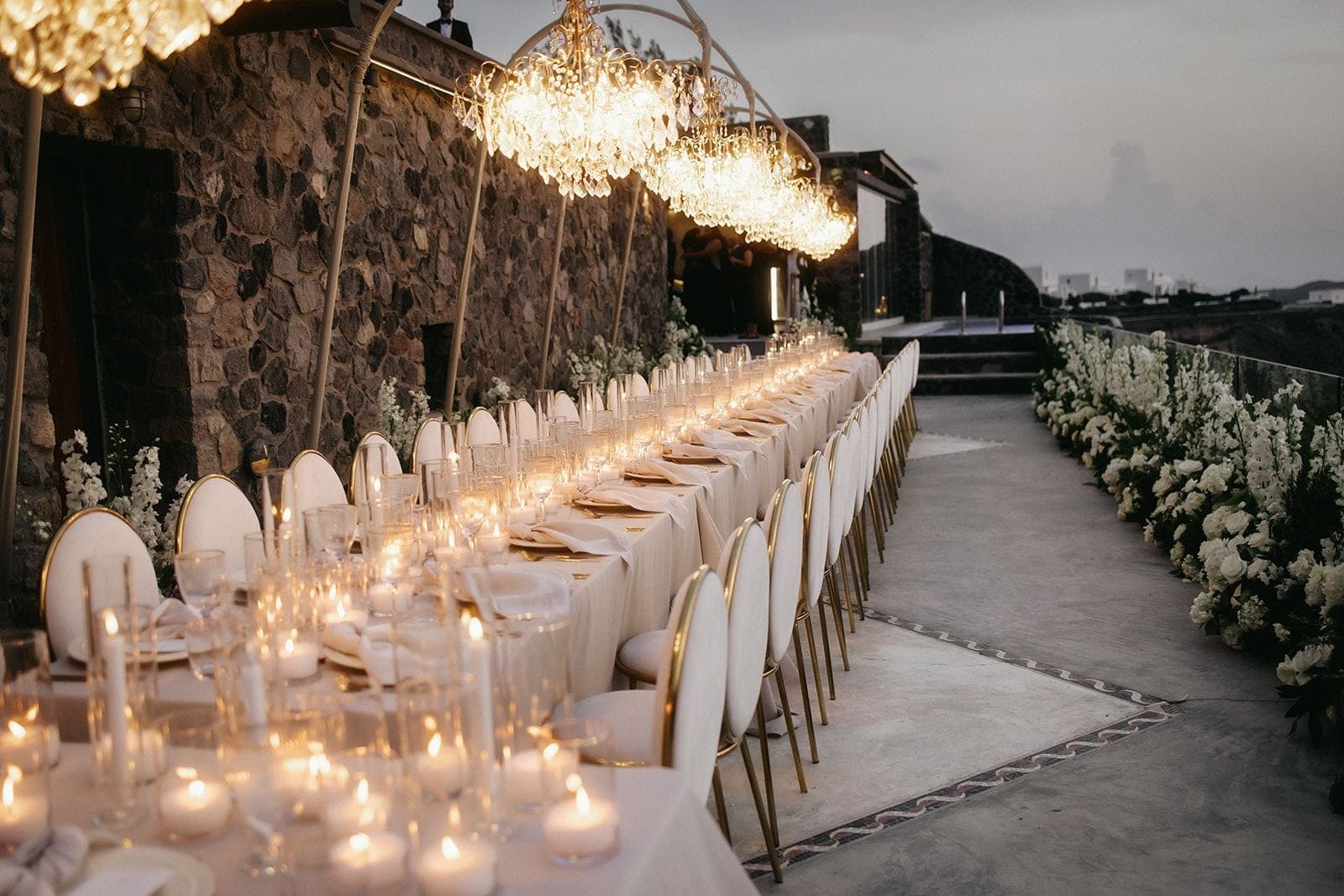 A long wedding dining table stretches across a terrace at Cavo Ventus Luxury Villa in Santorini, Greece, set with cream linens, gold-rimmed white chairs, and clusters of candles and clear glassware. Crystal chandeliers hang from gold arches above, and along the right edge a deep border of white stock and roses lines the path, with dark stone walls and a dusky evening sky behind.