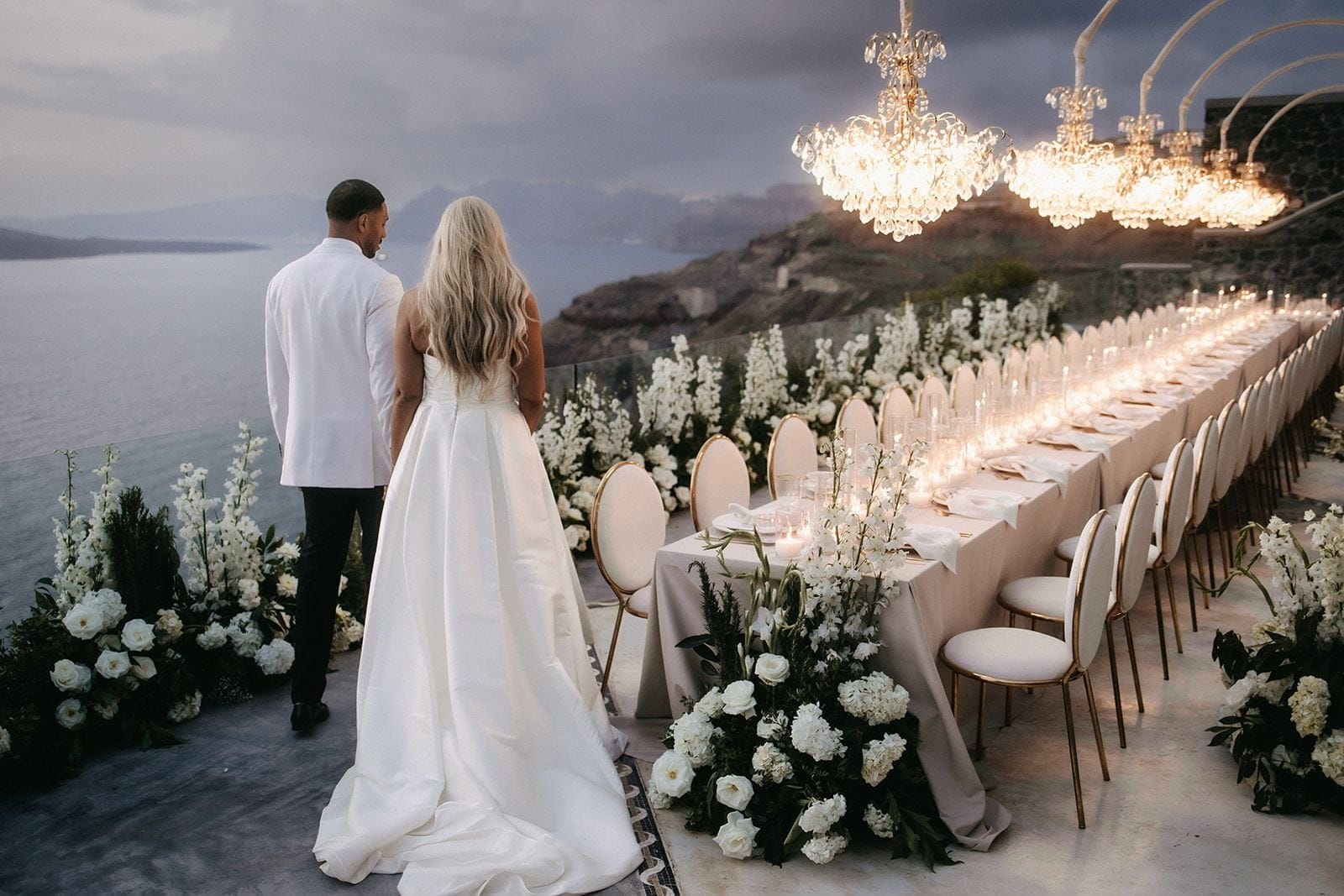 A bride with long, loose blonde waves in a strapless white ball gown and a groom in a white jacket and black trousers stand side by side, looking out over the sea from a clifftop terrace at Cavo Ventus Luxury Villa in Santorini, Greece. Beside them a long wedding table is laid with cream linens, gold-backed chairs and rows of candles, under crystal chandeliers, with bands of white roses, white delphinium and hydrangea arranged along its edges. It is dusk, with soft grey clouds and the lights glowing against the sea.