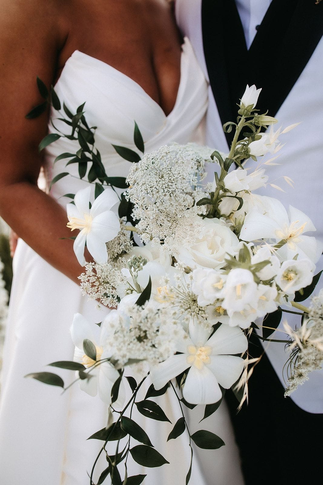 A cascading white wedding bouquet held against a bride's white strapless gown and a groom's black-lapelled white tuxedo, photographed close up at Cavo Ventus Luxury Villa in Santorini, Greece. The bouquet is loose and airy, with white lily-like blooms, creamy garden roses, delicate bell-shaped flowers, and frothy white Queen Anne's lace, all trailing green foliage and slender wheat-like stems. The soft, natural light and muted palette give it a calm, romantic feel.