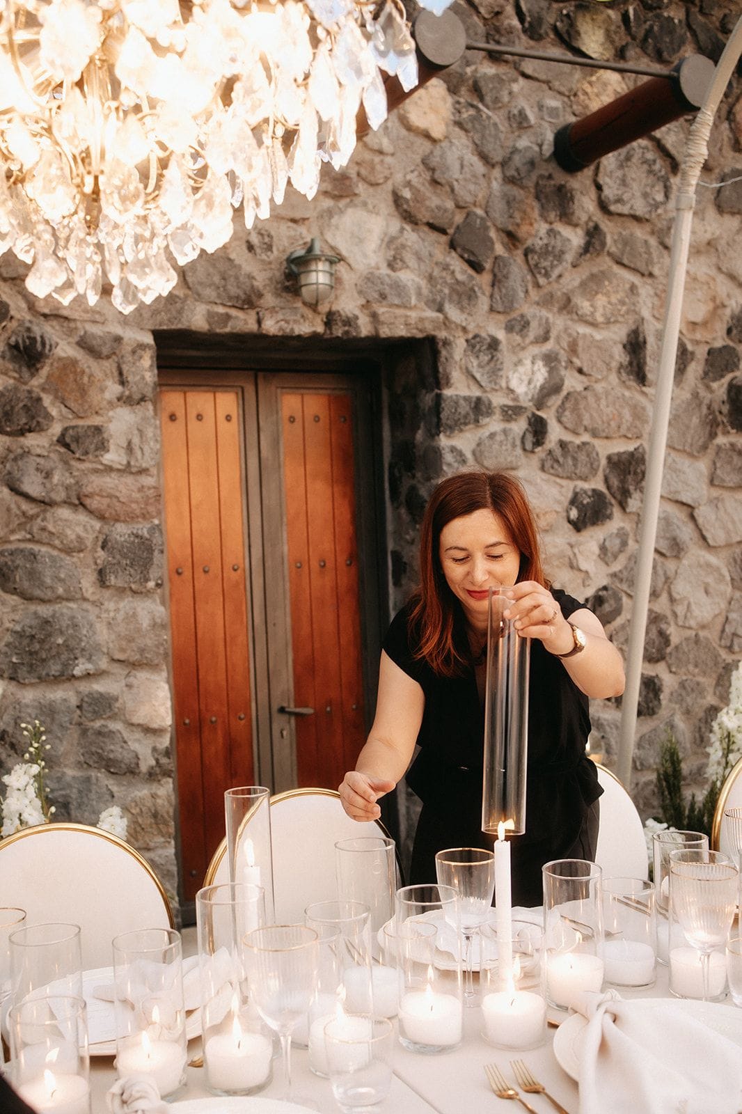 A woman with shoulder-length auburn hair, dressed in a black top and wearing a gold wristwatch, lowers a tall glass hurricane over a white pillar candle on a dinner table set for a wedding at Cavo Ventus Luxury Villa in Santorini, Greece. Rows of glass votives and pillar candles glow along the table beneath a crystal chandelier, with white cushioned chairs trimmed in gold and sprigs of white flowers and greenery at the table's edge. Behind her, a rough stone wall frames a pair of wooden doors, and the warm evening light gives the scene a calm, intimate mood.