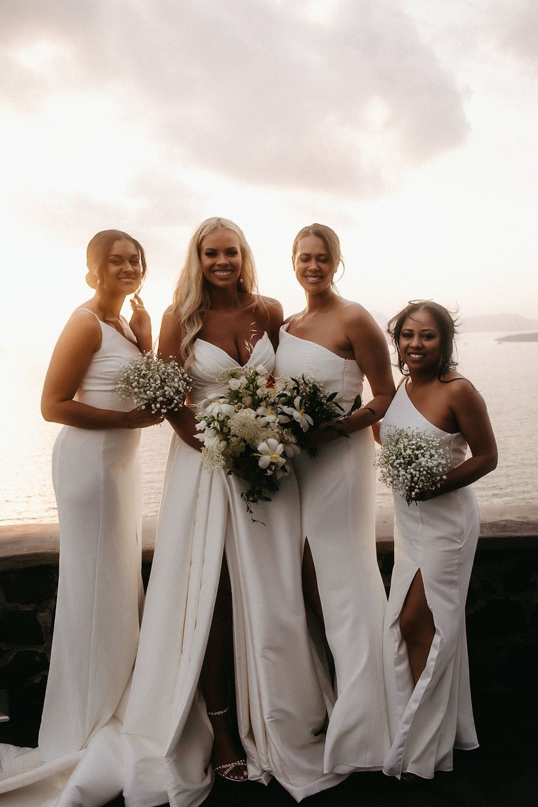 Four women in white satin gowns with side slits stand close together on a stone terrace at Cavo Ventus Luxury Villa in Santorini, Greece, with the sea glowing pale gold behind them at sunset. The bride, in the centre with long wavy blonde hair, holds a loose bridal bouquet of white blooms and dark green foliage, while the three bridesmaids each carry small posies of baby's breath. The soft, hazy light and wide, cloud-streaked sky give the scene a calm, romantic mood.