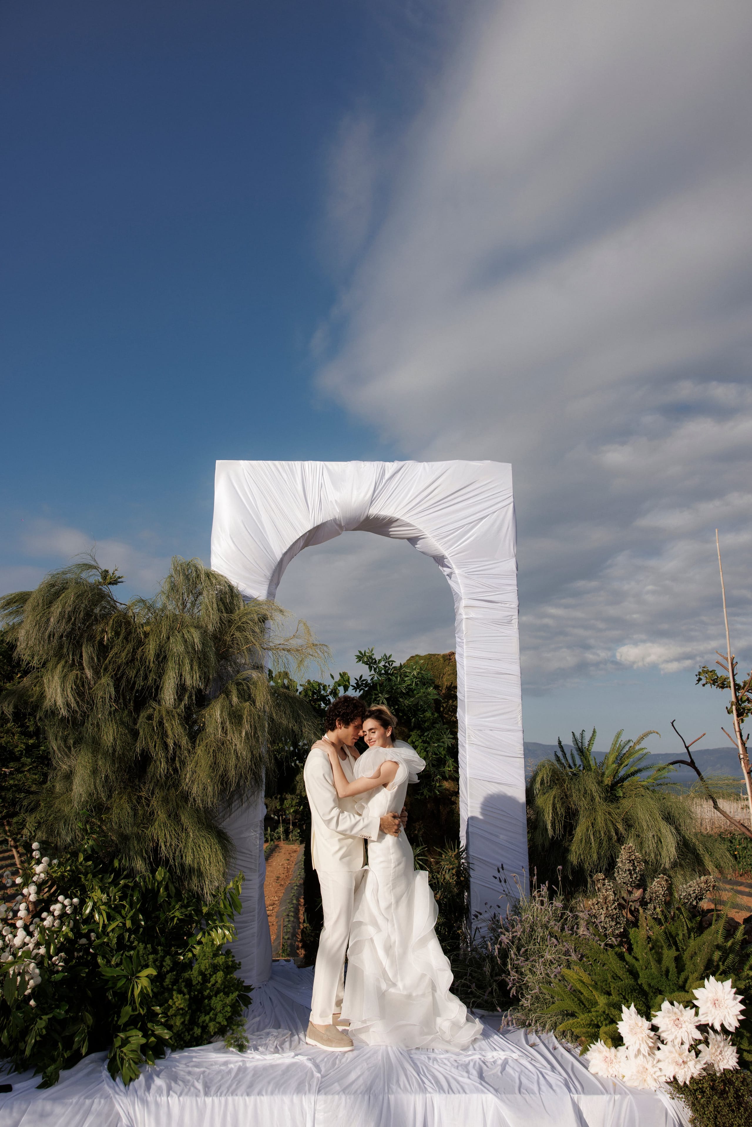 A bride and groom embrace closely on a white draped platform beneath a tall, white fabric arch at a wedding at Gramvousa Restaurant in Greece. He wears a cream suit and suede shoes, and she wears a white gown with ruffled organza skirts and a soft puffed sleeve, her eyes closed as she leans into him. Surrounding them are green foliage, feathery grasses, and cream dahlias in the foreground, with a hillside and distant sea under a soft blue sky.