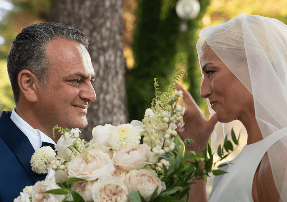A bridal bouquet of soft blush garden roses, white dahlias and sprigs of astilbe and white berries, wedding flowers at Sapel Hall in Greece. In the foreground, the bride in a white veil and sleeveless gown lifts a hand toward her smiling partner, who wears a navy suit, as they gaze at each other beneath a tree in warm, dappled sunlight.