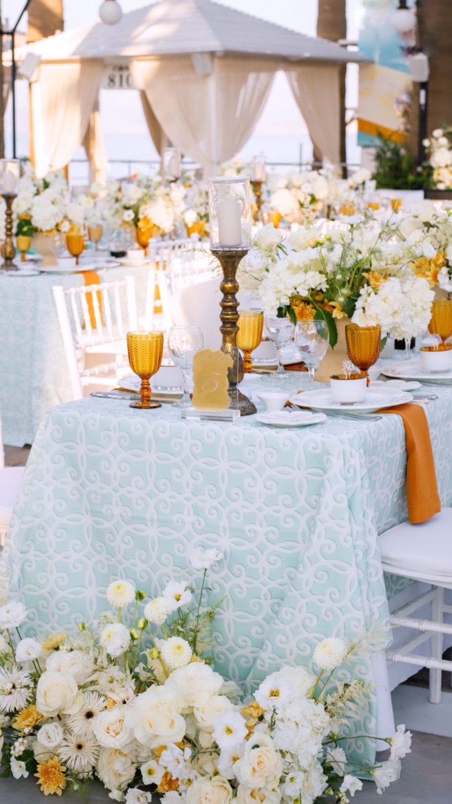 A long wedding table dressed in a pale mint tablecloth with white scrollwork, set with amber goblets, white plates and burnt-orange napkins under a white gazebo with billowing cream drapes at Sapel Hall in Agia Marina, Crete, Greece. Clusters of white hydrangeas and yellow blooms rise from the table, and a cascade of white roses, ranunculus and marigold-yellow flowers spills over the floor at the table's base, with a brass candleholder holding a tall pillar candle at its centre.