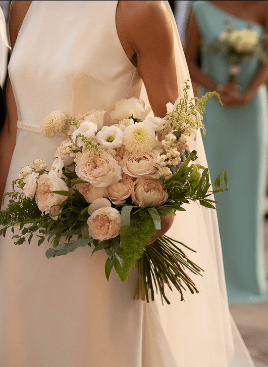 A bride in a simple white satin gown holds a loose, romantic bridal bouquet of blush garden roses, white ranunculus-style blooms, and small cream lisianthus, with trailing green foliage and sprigs of astilbe. Her dress and the bridesmaids' pale blue-green gowns behind her suggest a warm outdoor ceremony at SAPEL Hall in Agia Marina, Crete, Greece.
