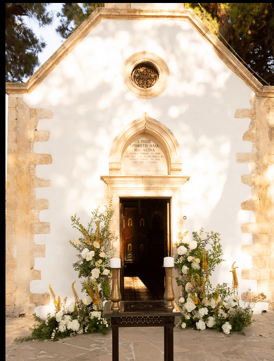 Two tall arrangements of white flowers, likely garden roses and hydrangea, with trailing green foliage and spikes of yellow blooms, frame the open wooden doorway of a whitewashed stone chapel. Two tall candles stand on brass holders on a dark wooden stand in front of the door. This is a wedding flower display at Sapel Hall in Agia Marina, Crete, Greece, set in warm late-afternoon light.