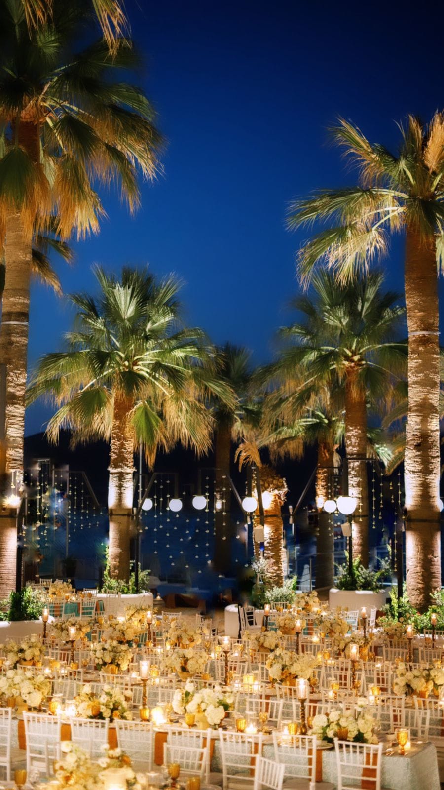 Rows of round tables under a dusk-blue sky, each dressed with white hydrangea-style blooms and low arrangements, glowing with candles in amber glass holders, at a wedding reception in Greece. Tall, uplit palm trees frame the scene, and strings of soft white lights hang between them over the terrace at SAPEL HALL in Agia Marina, Crete.
