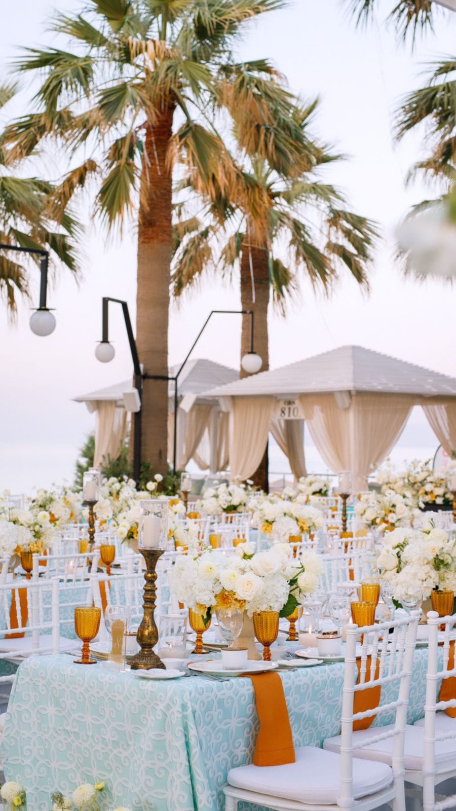 Long banquet tables draped in pale aqua damask sit under tall palm trees at a seaside wedding reception at Sapel Hall in Agia Marina, Crete, Greece. Each place setting has amber goblets, burnt-orange napkins and white chiavari chairs, with low arrangements of white garden roses, hydrangeas and touches of golden yellow blooms. Cream-curtained cabanas and tall black lamps frame the soft pinkish evening sky beyond.