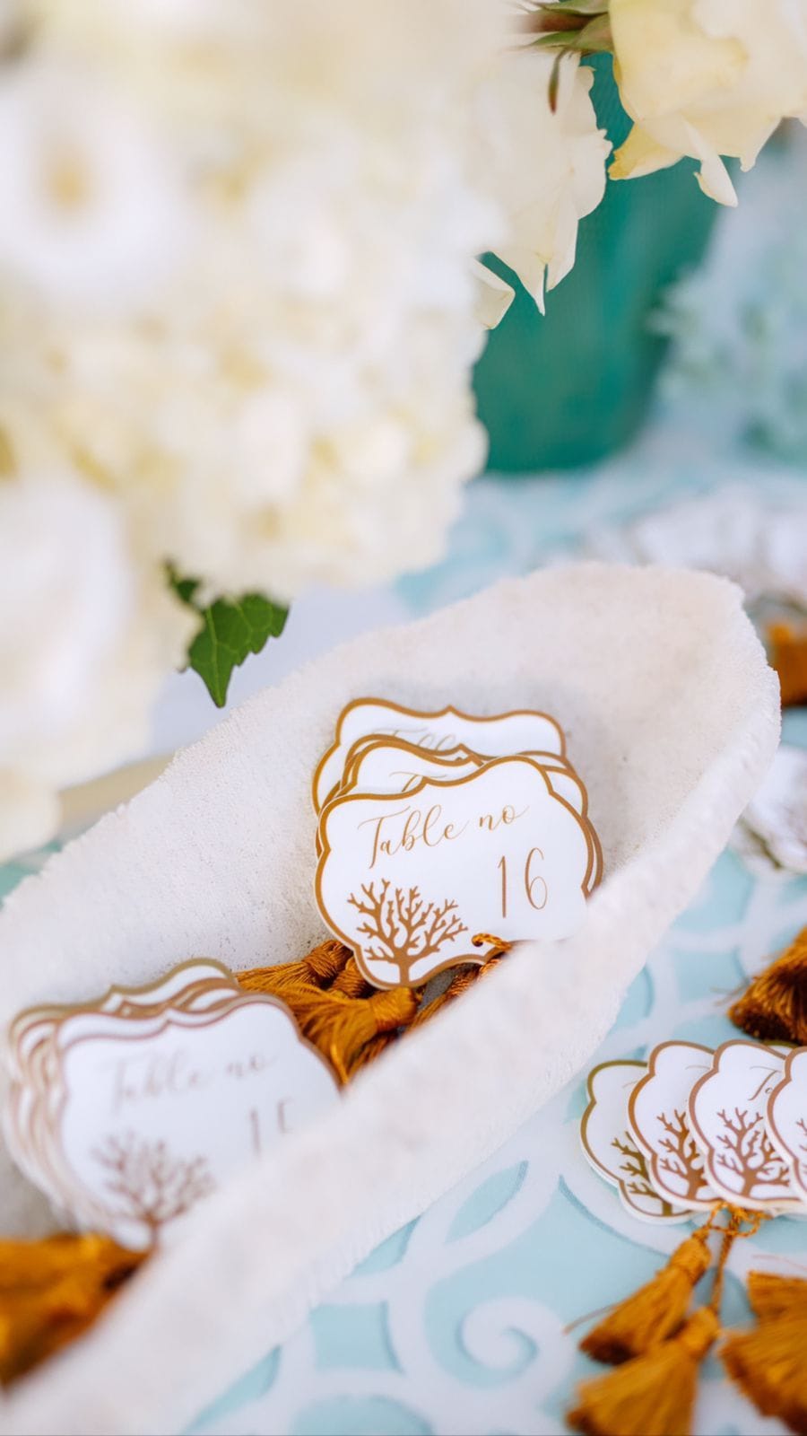 A wedding table setting at Sapel Hall in Agia Marina, Crete, Greece, with white hydrangeas in the soft-focus background and a teal vase beside them. In the foreground, a cream sponge-like dish holds several white table number cards edged in gold, with a tree motif and "Table no 16" in gold script, each cards tied with a gold tassel. The pale teal tablecloth with a white pattern completes a calm, elegant palette.