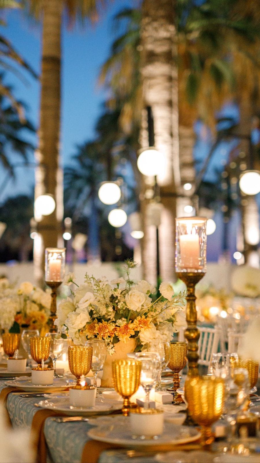 A long wedding dinner table at dusk at Sapel Hall in Agia Marina, Crete, Greece, set with white roses, cream hydrangeas and orange chrysanthemums in low centrepiece vases. Tall gold candlesticks hold glowing pillar candles, and amber goblets sit beside clear glassware on pale blue linens. Behind the table, tall palm trees and round white lanterns glow against the deep blue evening sky, creating a warm, golden mood.