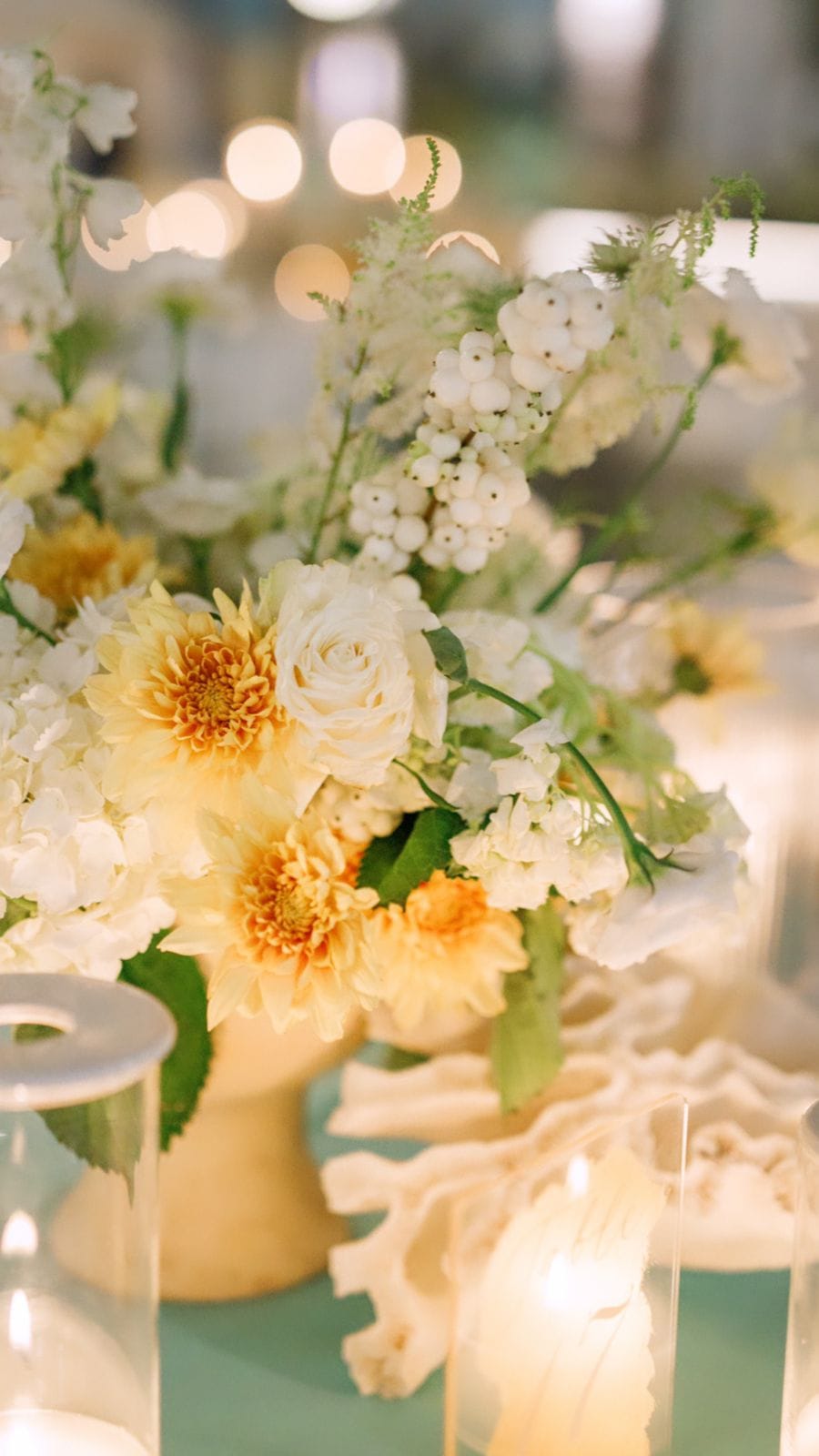 A wedding table centrepiece at Sapel Hall in Agia Marina, Crete, Greece, built around soft cream and butter-yellow blooms: a pale garden rose, yellow dahlias and white hydrangea, with clusters of small white berries and feathery white sprays rising above. The stems sit in a cream footed vase on a pale sea-green cloth, flanked by glass hurricane candles glowing warmly, with white coral pieces scattered nearby and blurred golden lights in the background.