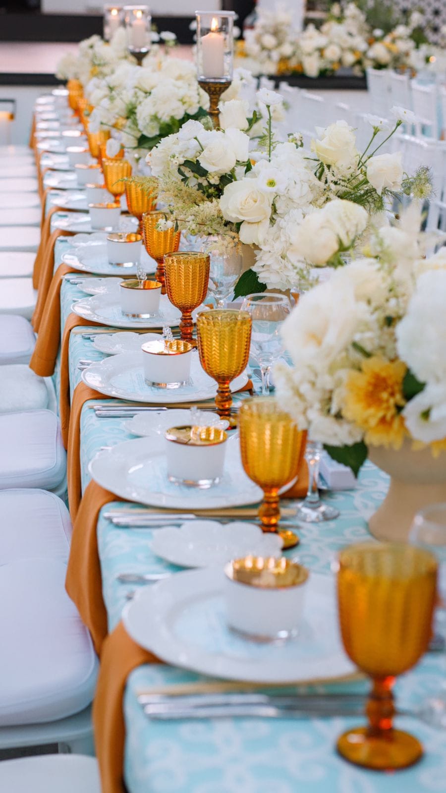 A long banquet table laid for a wedding dinner at Sapel Hall in Agia Marina, Crete, Greece, with white plates, gold-rimmed candle cups and amber goblets set on pale turquoise linen. Along its length rise arrangements of white garden roses, hydrangeas and feathery white filler, with a few golden chrysanthemums tucked in, and tall glass hurricane candles glow at the back. Empty white chairs line the room, and the soft, even light gives the scene a calm, elegant feel.