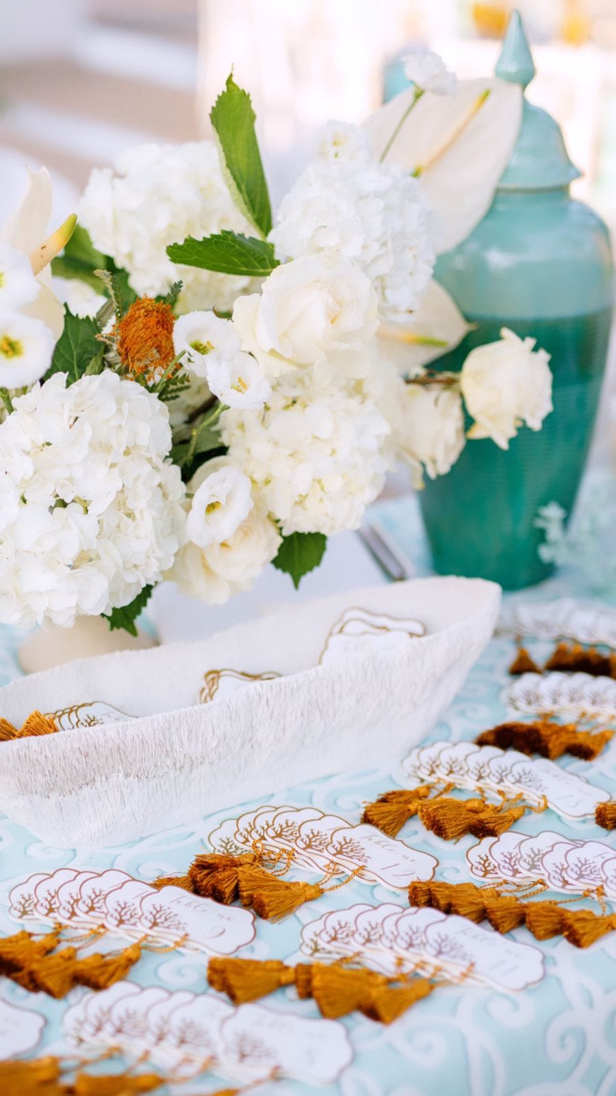 A wedding table at Sapel Hall in Agia Marina, Crete, Greece, dressed with a loose arrangement of white hydrangeas, white roses and calla lilies with green leaves and a touch of orange banksia. Beside it sits a white woven basket, and on a pale blue patterned cloth lie place cards with gold-edged tassels in ochre, while a turquoise ceramic urn adds a cool note to the warm, sunlit scene.