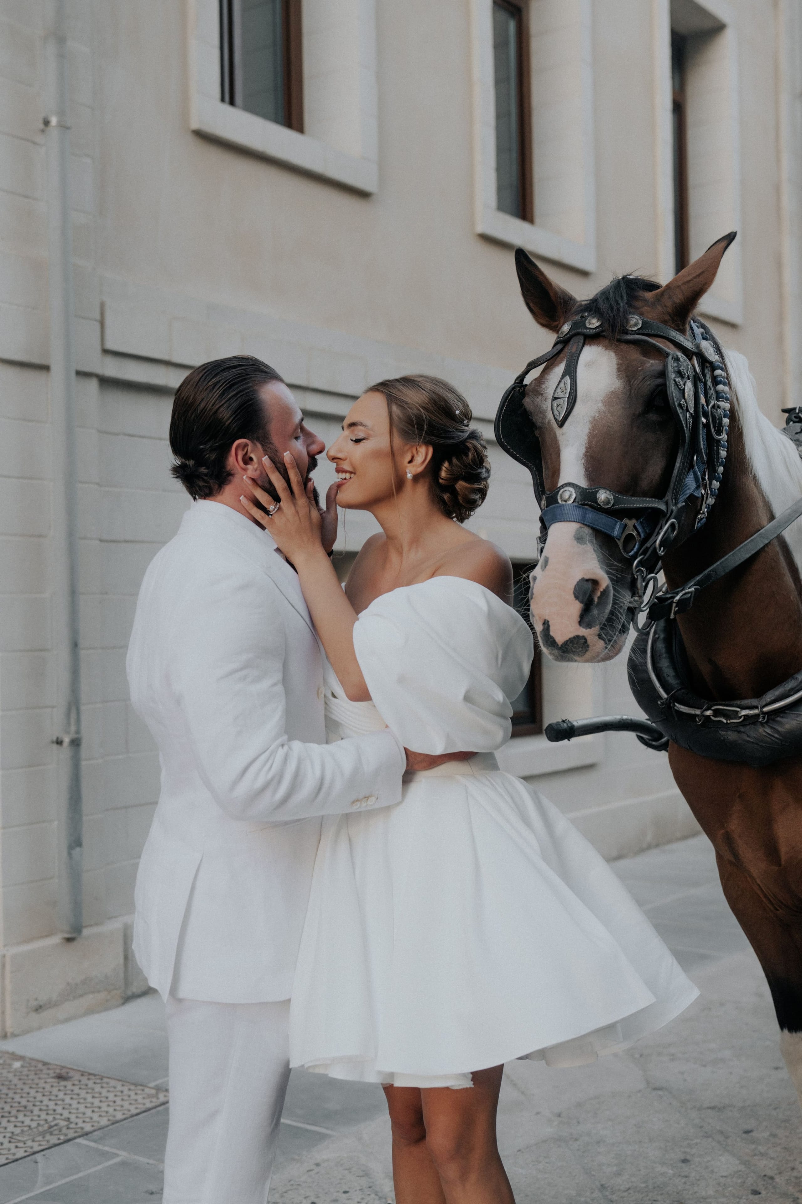 A bride in an off-the-shoulder white gown with a sculpted sleeve and a soft updo holds her groom's face in her hands as they share a tender moment, both smiling, while a brown horse with a white blaze and a dark ornate bridle stands close beside them against a pale stone wall. This is a wedding photograph taken in Greece, with no flower arrangements visible in the frame.