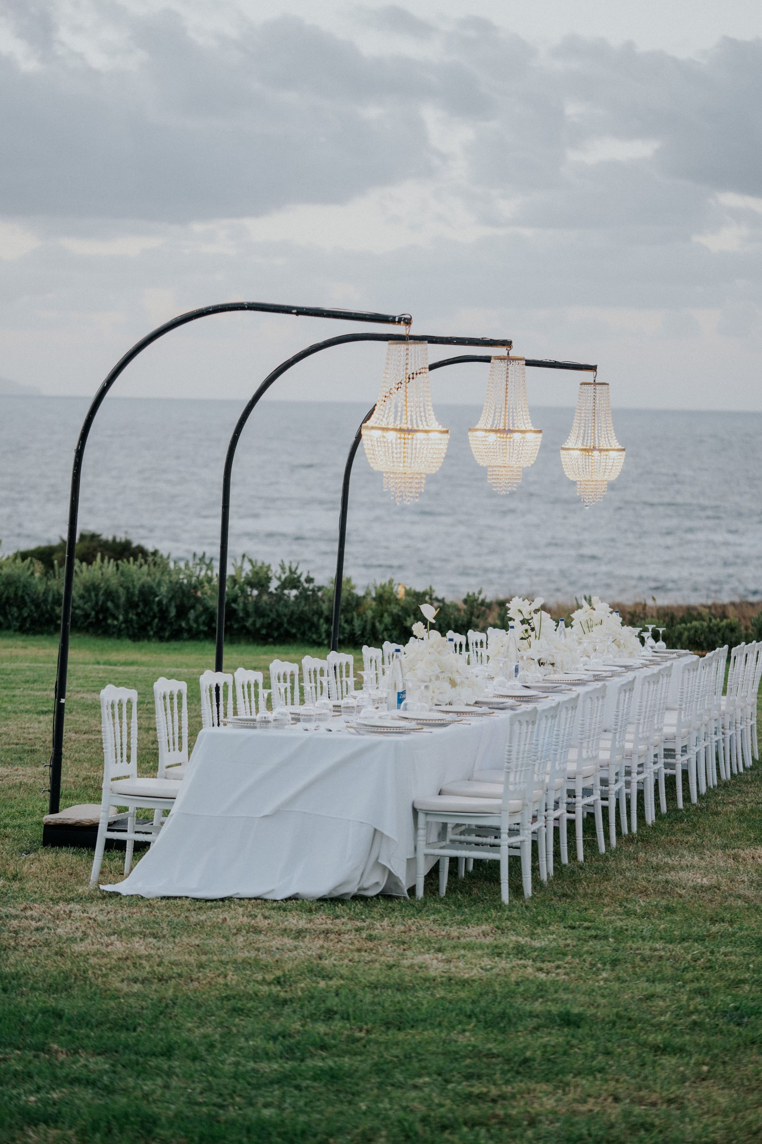 A long wedding dining table on a lawn above the sea in Greece, dressed in a white linen cloth and lined with white chiavari-style chairs. Three black arched frames hang crystal chandeliers over the table, and low centrepieces of white roses and white orchids sit along its length. The flat blue-grey sea and a hedge of green shrubs fill the background under a pale, soft evening sky.