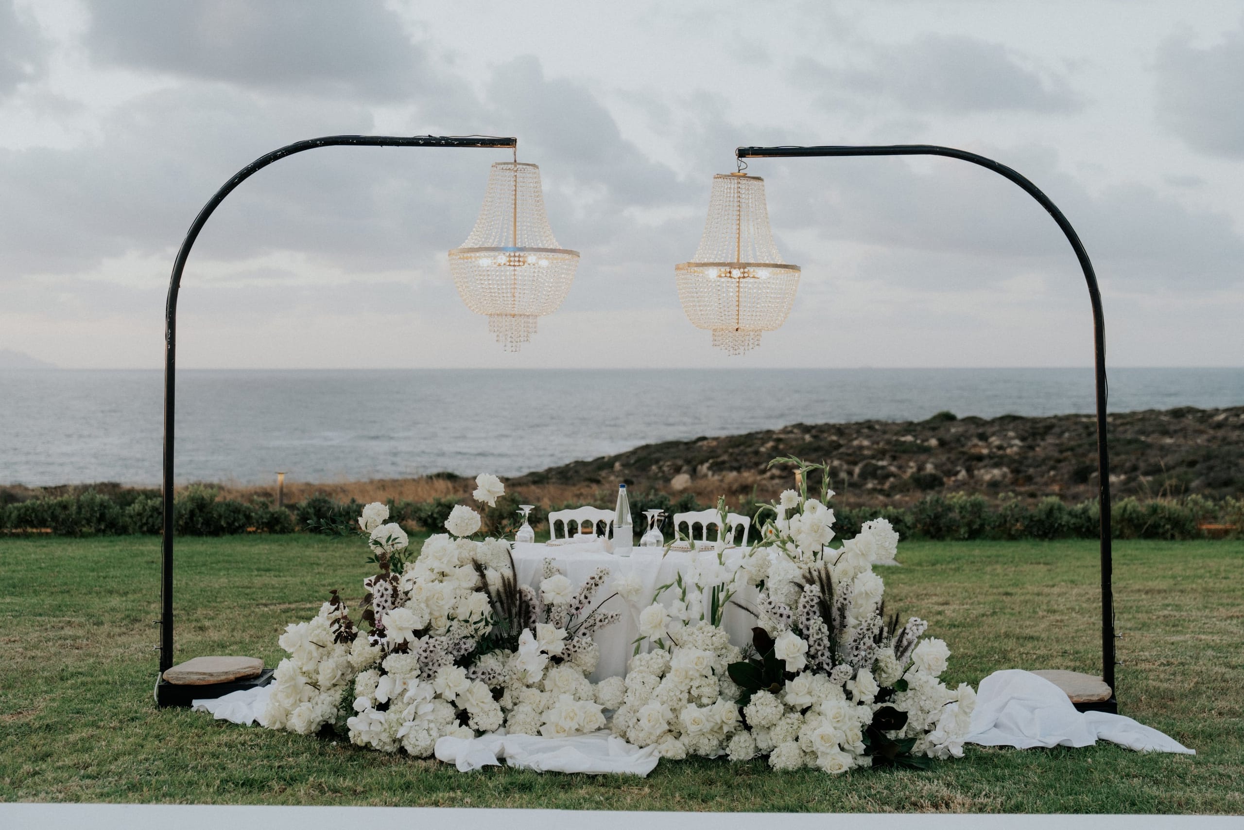 A lush all-white sweetheart table on a grassy clifftop, its base heaped with white roses, hydrangeas and gladioli dotted with feathery grasses and dark foliage. Two crystal chandeliers hang from black arched frames above it, with the calm grey sea and rocky shoreline behind, under a soft overcast sky. This is a wedding table flower display in Greece.
