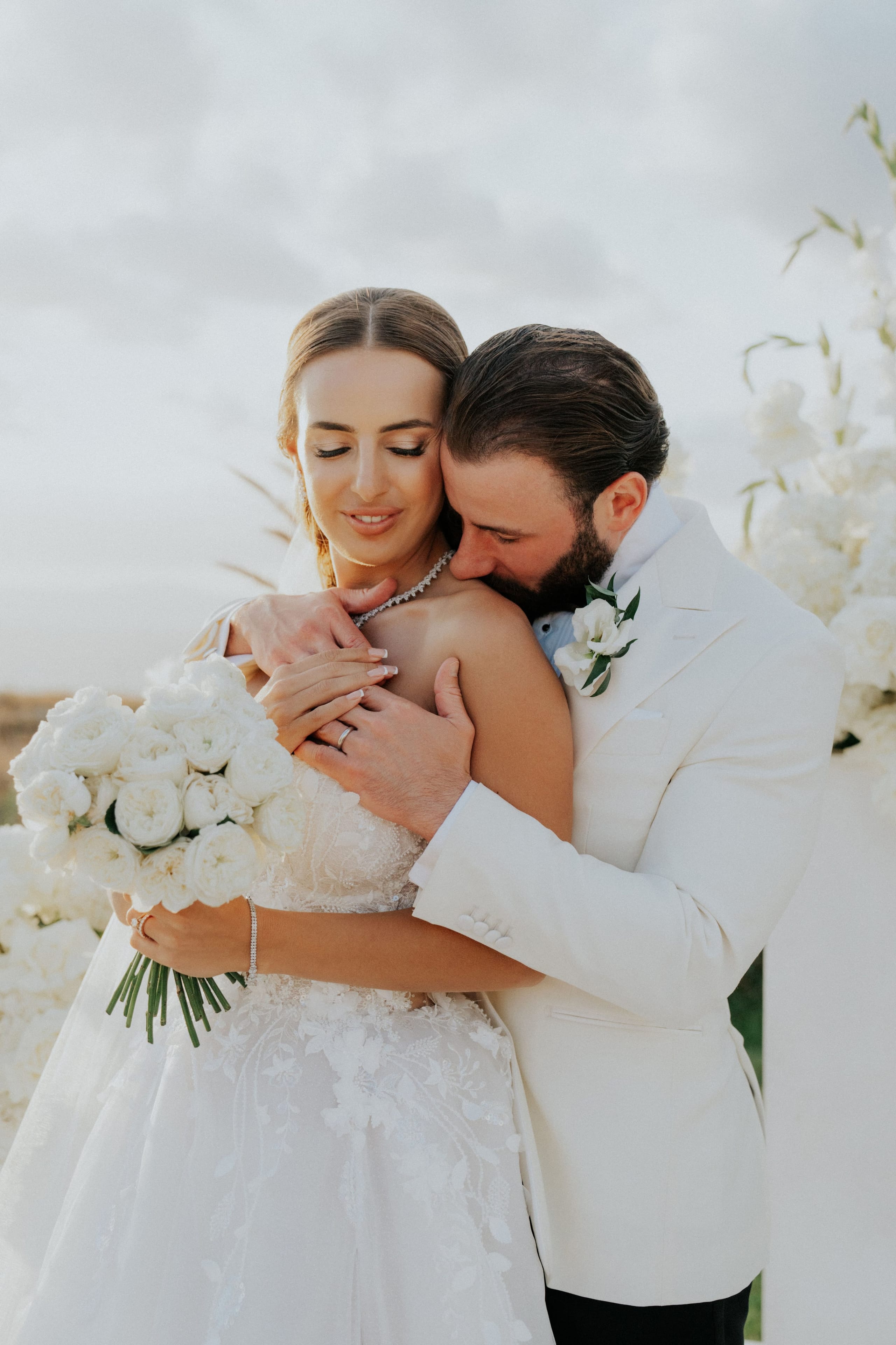 A bride with her hair swept back and eyes lowered, smiling softly, as her groom in a cream suit with a white bloom boutonniere leans in to rest his head against her shoulder. She holds a dense bouquet of white garden roses, and a white floral installation of similar blooms fills the background against a pale, overcast sky. This wedding moment, photographed in Greece, is all soft whites and gentle light.