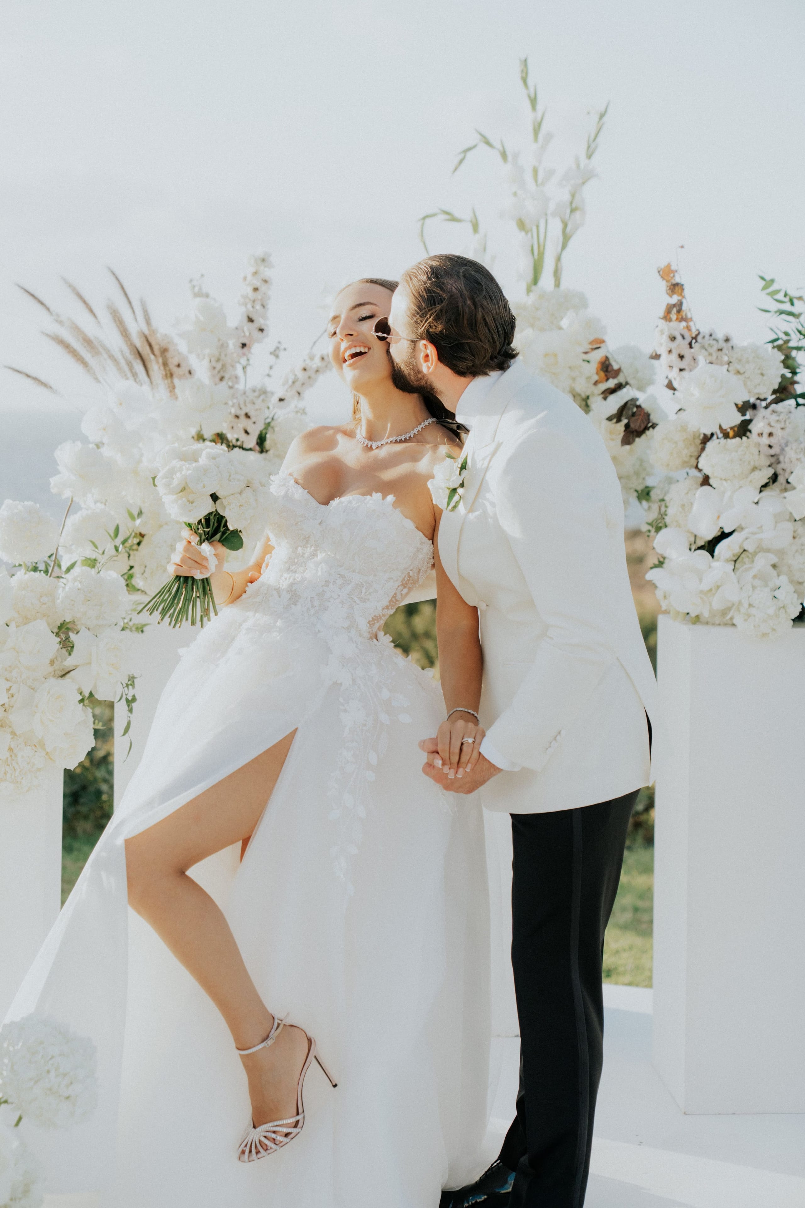 A bride in a strapless white gown with a sculpted lace bodice and a slit to the thigh holds a loose bouquet of white roses and hydrangeas, while a groom in a white dinner jacket and black trousers leans in close beside her. Their hands are clasped at the centre, and behind them a white arch is dense with white hydrangeas, roses and foliage, set at a wedding in Greece.