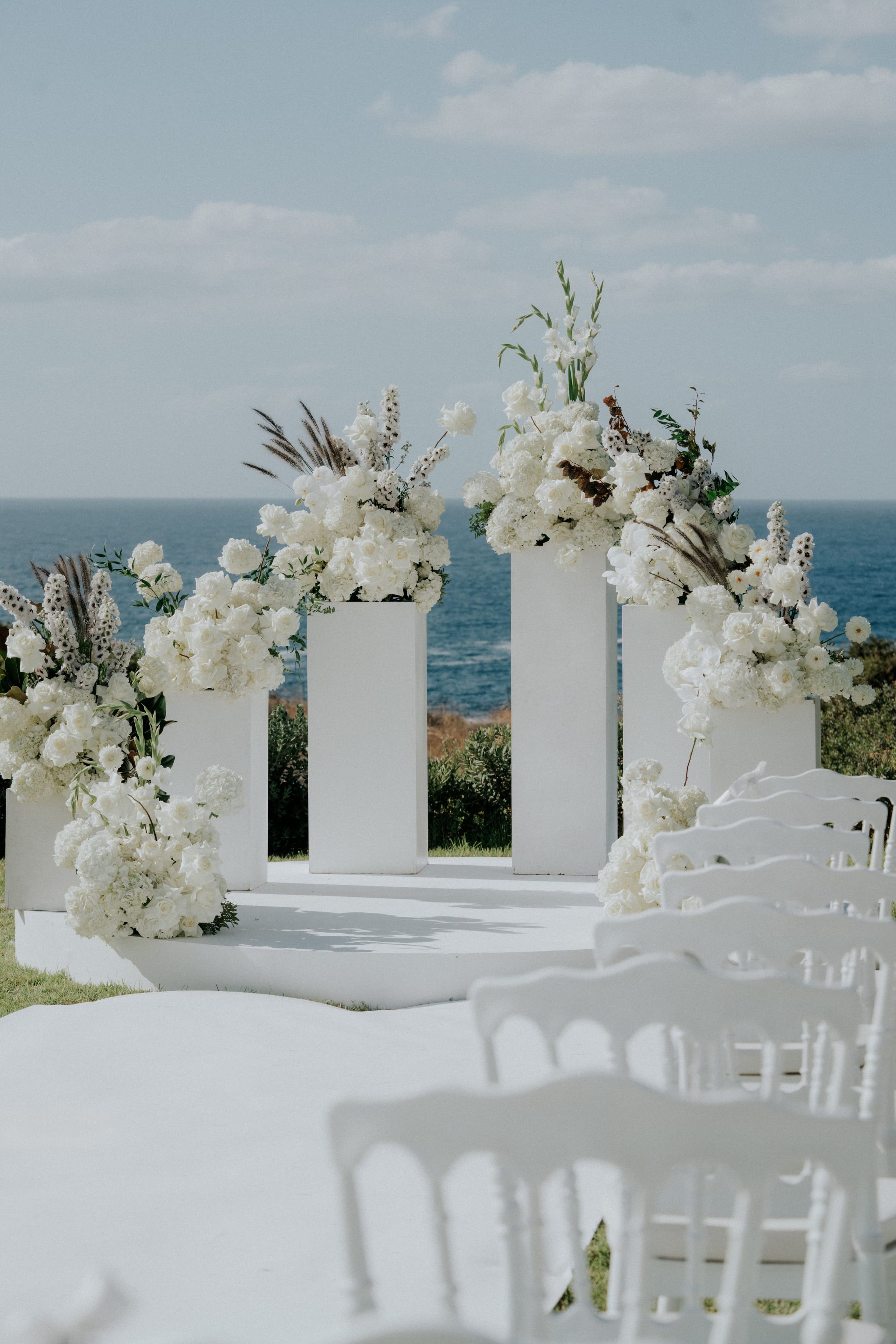 A wedding ceremony setup in Greece where four tall white pedestals, each crowned with dense white arrangements of roses, hydrangeas and delphinium, stand on a white circular platform above a deep blue sea. Feathery plumes and dried grasses add texture to the tops, and rows of white chairs lead toward the stage in the foreground. The bright sunlight and the calm horizon give the scene a fresh, serene feel.