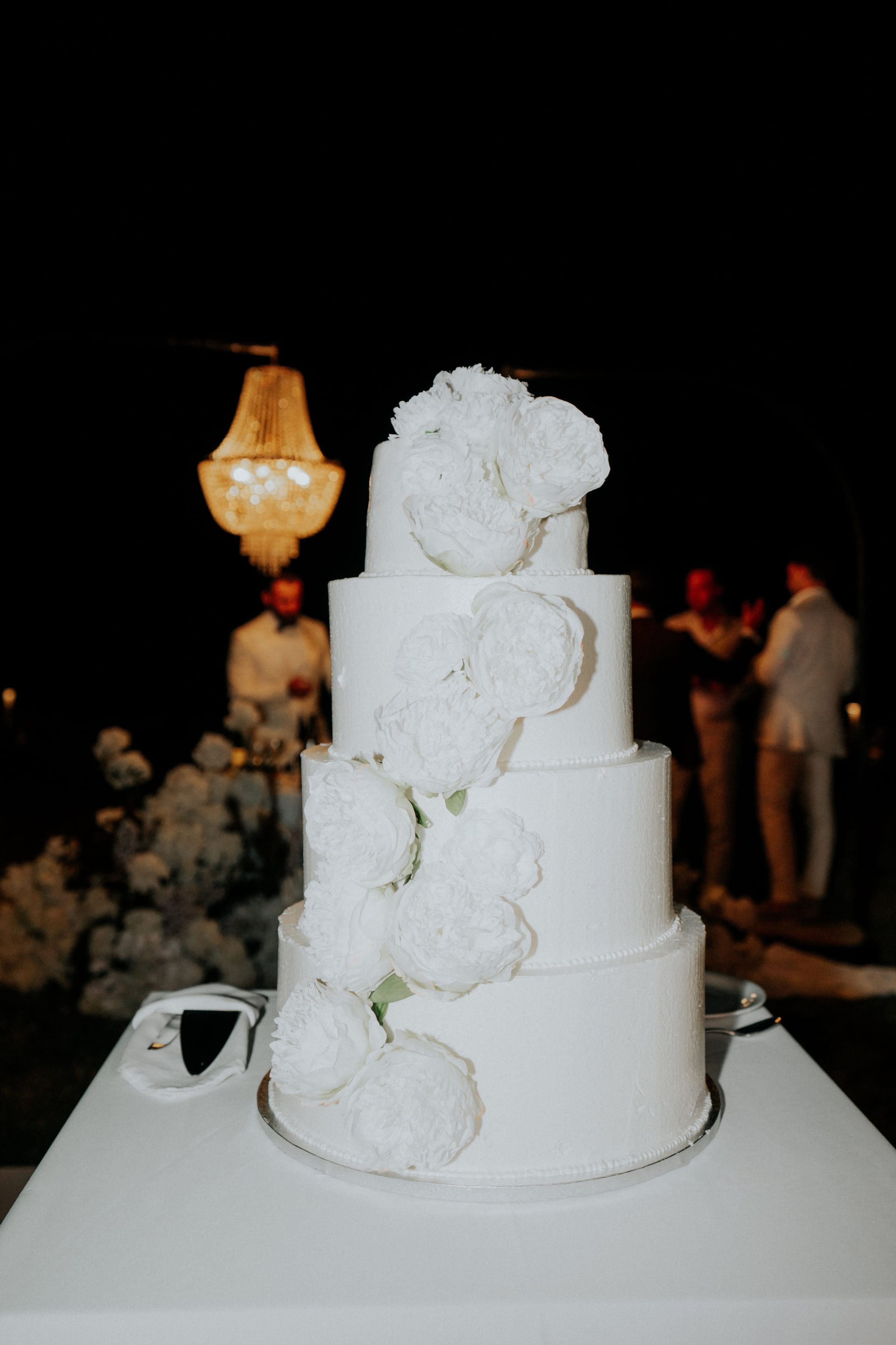 A four-tier white wedding cake with smooth buttercream sides and a cascade of large white peonies spiralling up from the base to the top, the blooms looking like sugar or fabric. It stands on a table at night at a wedding in Greece, with a glittering crystal chandelier glowing softly behind it and guests in pale suits blurred in the background.