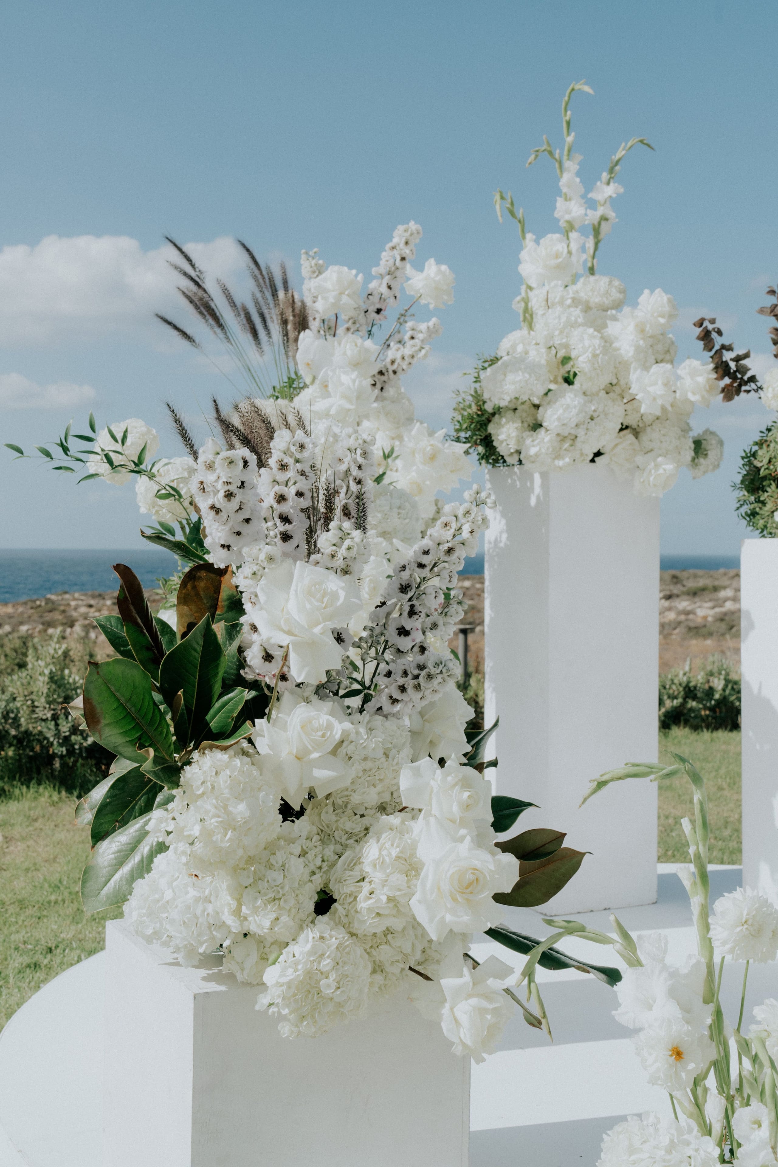 A tall, lush arrangement of all-white wedding flowers sits on a white plinth, with white garden roses, hydrangeas and spires of white delphinium dotted with dark centres, set against dark magnolia leaves and feathery grass plumes. Behind it, more white blooms crown a pedestal, and the calm blue sea and rocky shoreline of Greece stretch out under a pale sky.