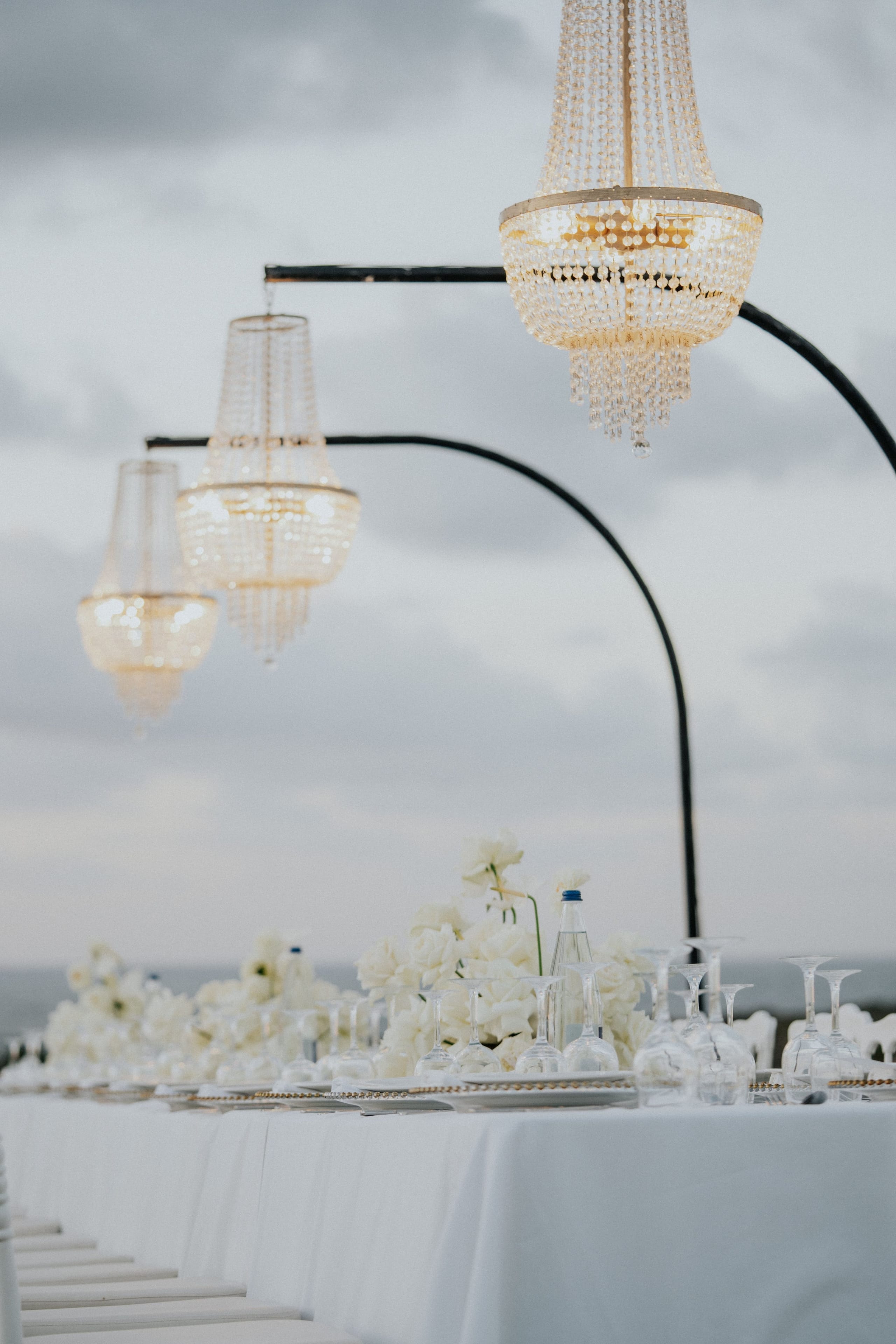Three crystal-beaded chandeliers hang from curved black metal arms against a soft, overcast sky, their glass strands catching the light at different heights. Below, at the bottom edge of the frame, the tops of white flowers and a glass bottle sit on a table, hinting at a wedding table setting in Greece. The muted grey and cream palette gives the scene an airy, elegant mood.