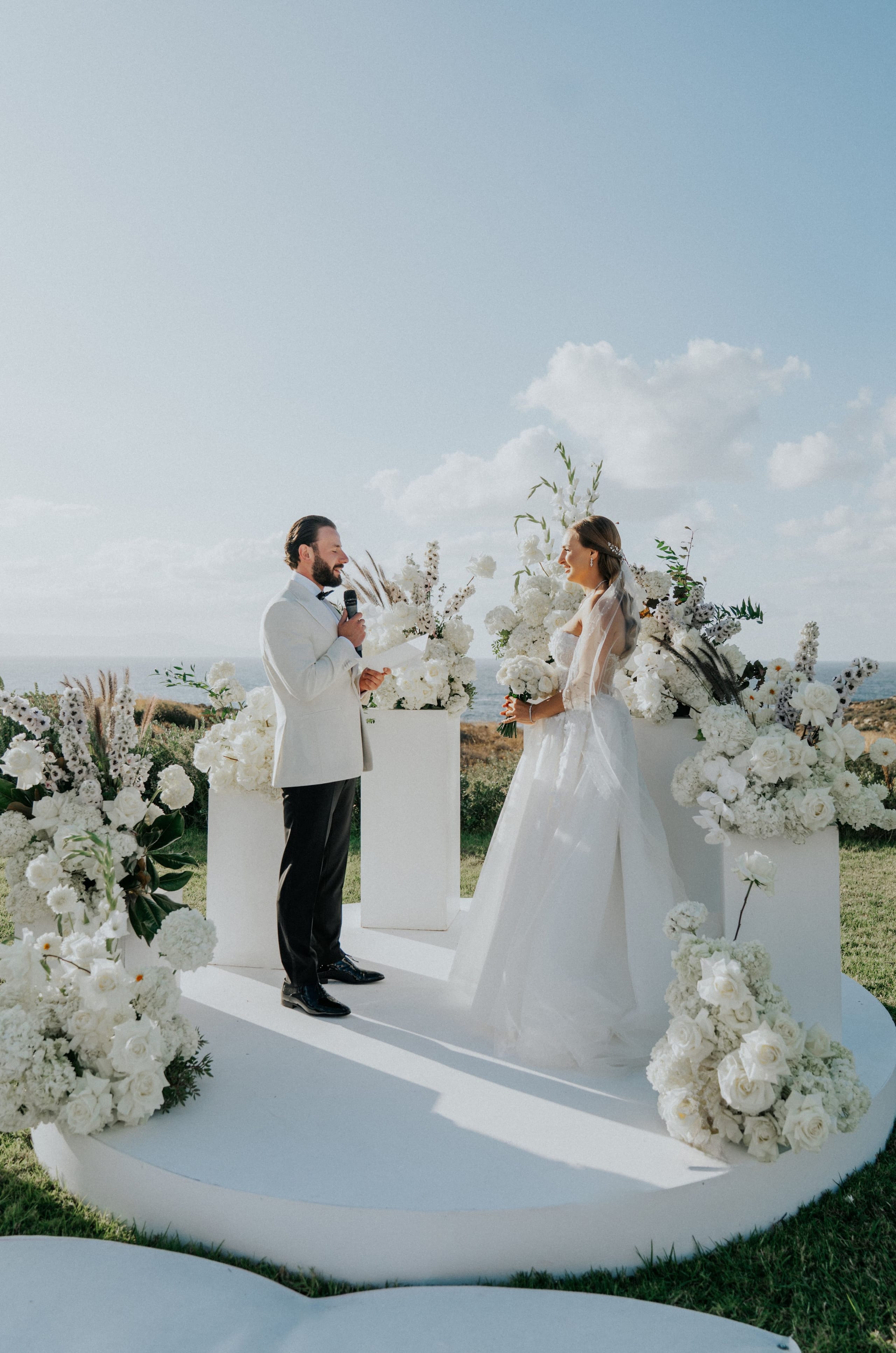 A bearded groom in a cream jacket and dark trousers holds a microphone and speaks to his bride, who wears a strapless tulle gown and holds a white rose bouquet, as they stand on a round white platform at a wedding ceremony in Greece. White garden roses, hydrangeas and tall white delphinium spires with feathery grasses frame the platform, and the sea glimmers on the horizon behind them under soft afternoon light.