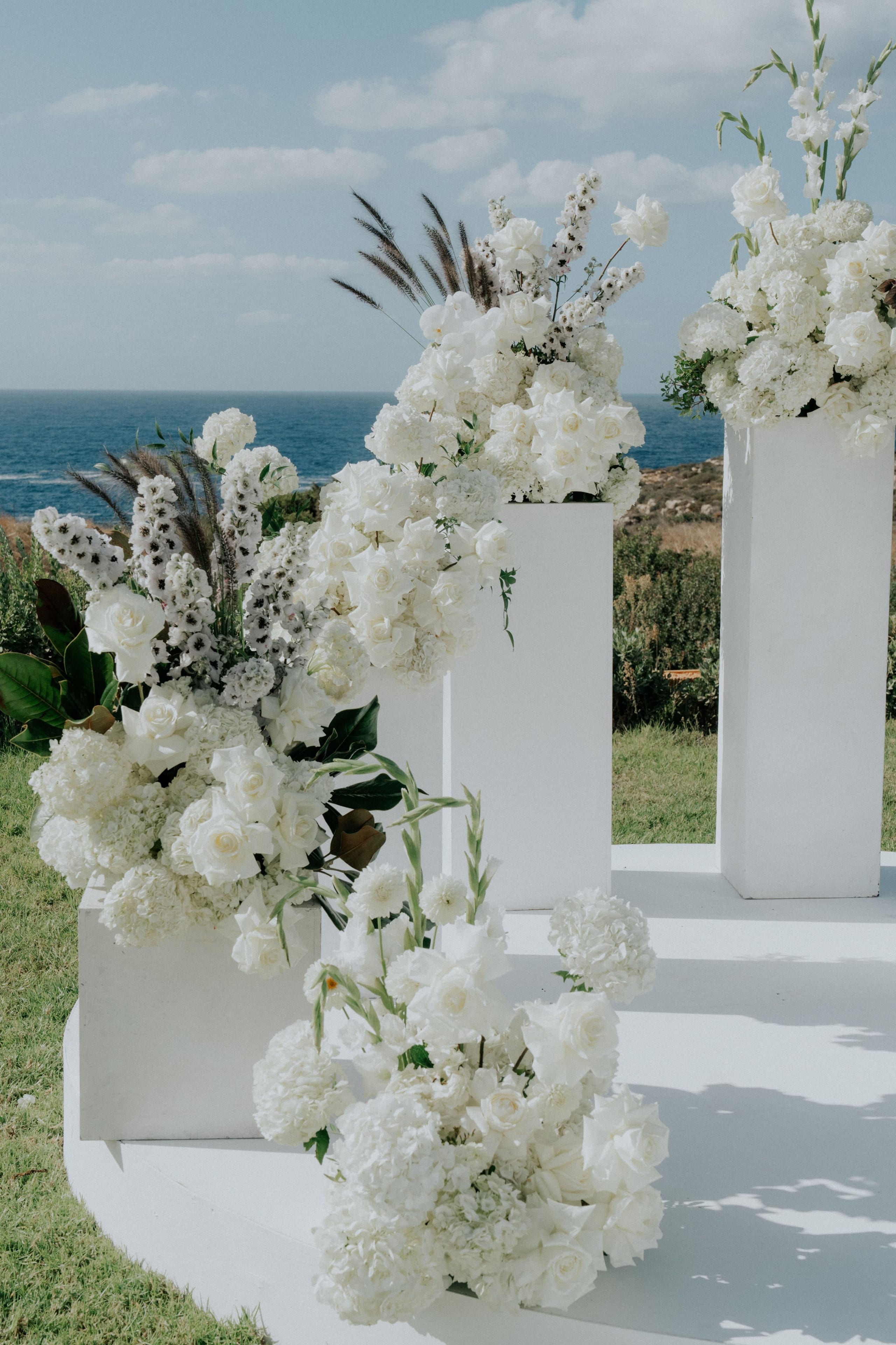 Three tall white pedestals on a white platform hold lush all-white arrangements of roses, hydrangeas and white delphinium, with dark pampas-like grasses and magnolia leaves adding depth, while a low spray of gladioli and white roses sits in the foreground. Behind them the deep blue Mediterranean sea and dry scrubby hillside frame a wedding ceremony setup in Greece.
