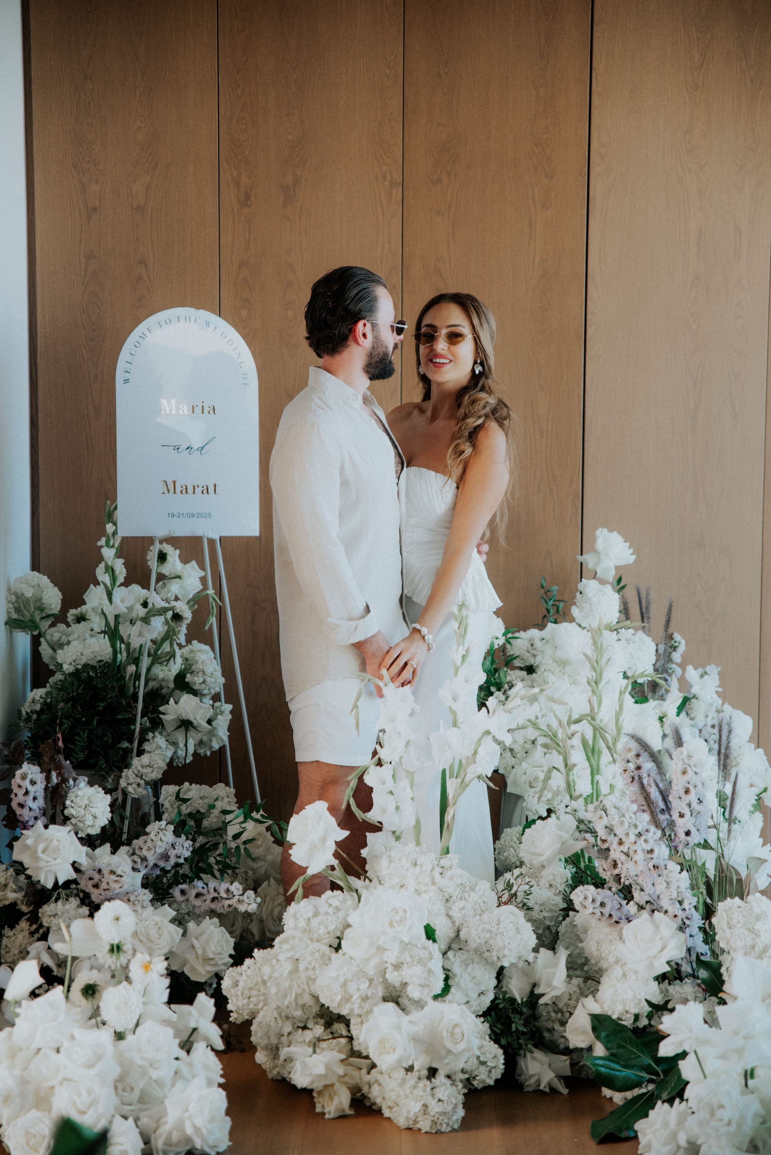 Dense, all-white floral installations fill the frame, with a couple standing among them from the waist down: he wears a cream linen suit and she a pale, pleated top and skirt, her hand resting in his. Creamy white garden roses, hydrangeas, gladioli, delphinium spires and sprigs of dark green foliage spill across the wooden floor against a warm timber wall, and a small sign reading 19-21/09/2025 stands on an easel at the left. This is a wedding flower display in Greece, styled in soft, monochrome whites.
