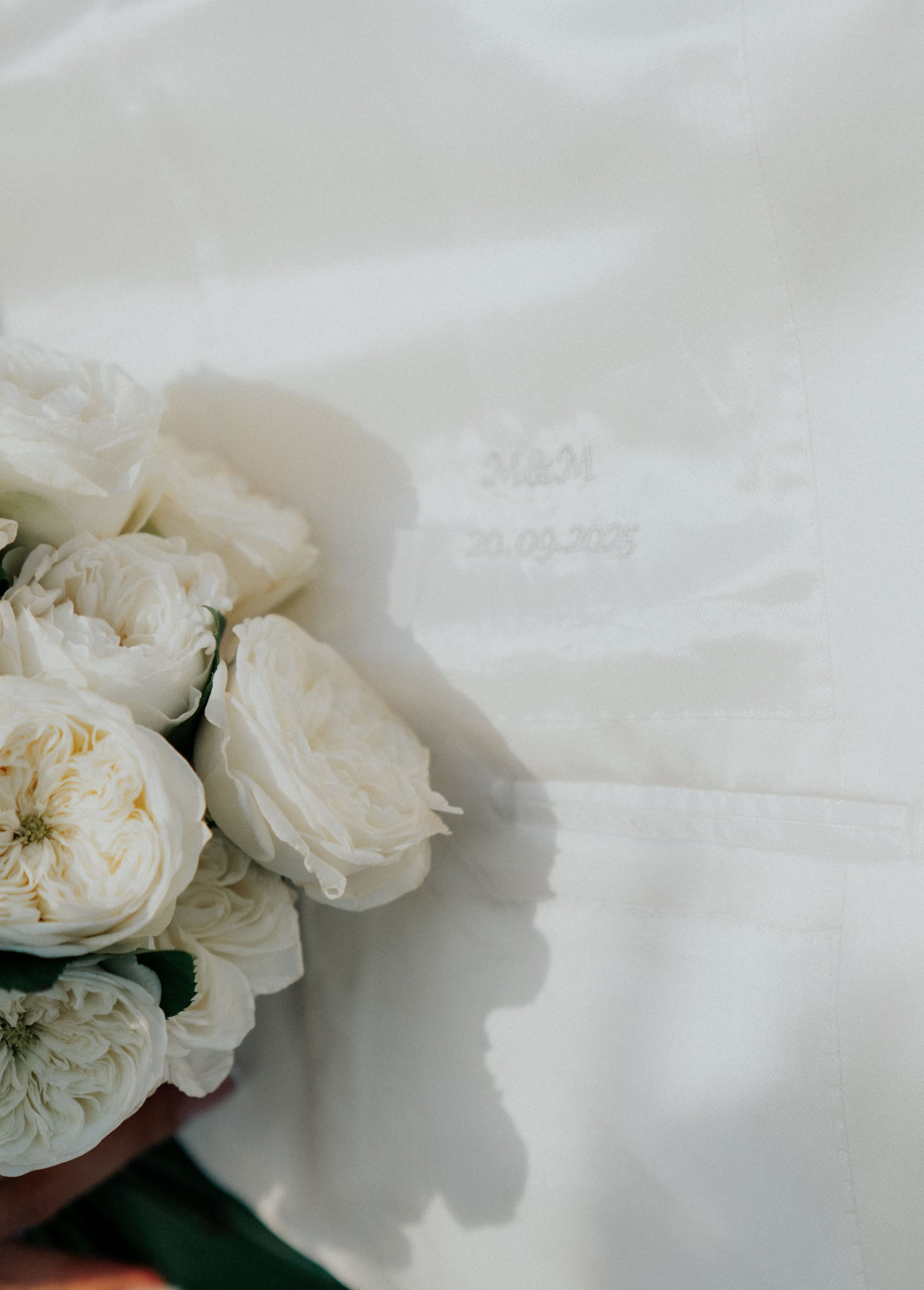 A close view of a bridal bouquet of creamy white garden roses with ruffled, layered petals, held at the left edge beside a crisp white fabric with a faint embroidered date, 20.09.2025, stitched in white thread. Dark green leaves peek out below the blooms, and soft shadows fall across the fabric in warm, natural light. This is a wedding flower detail from Greece.