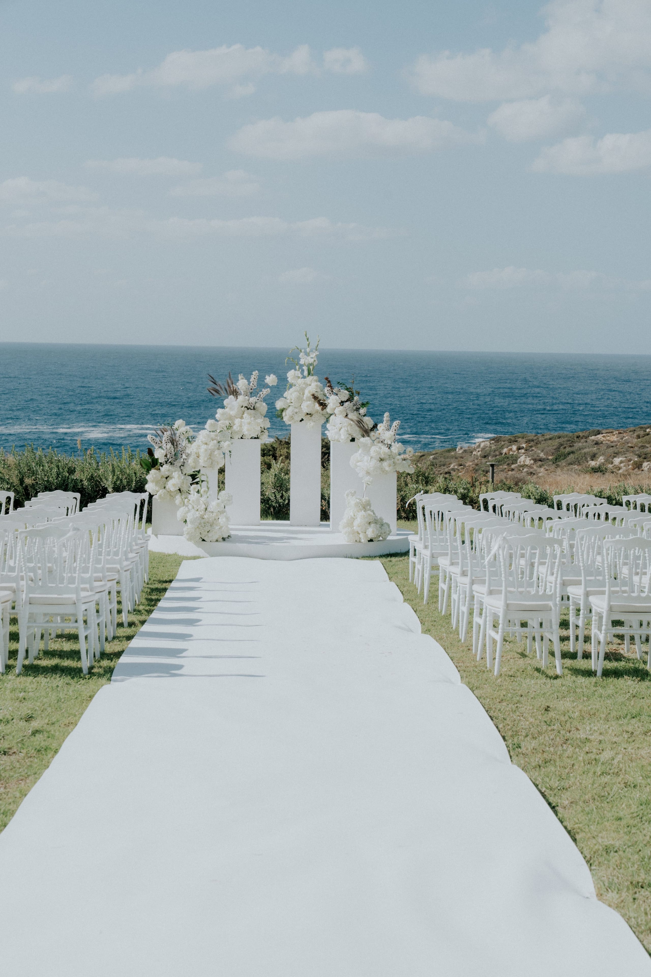A clifftop wedding ceremony set-up in Greece, with a white aisle runner leading between rows of white chiavari-style chairs to a round white platform. Three tall white pedestals stand on it, crowned with dense white blooms that look like hydrangeas and garden roses, with white gladioli and dried feathery grasses tucked among them. Beyond the flowers, the deep blue Aegean stretches to a pale horizon under a soft sky, with scrubby green shrubs and rocky slopes framing the scene.
