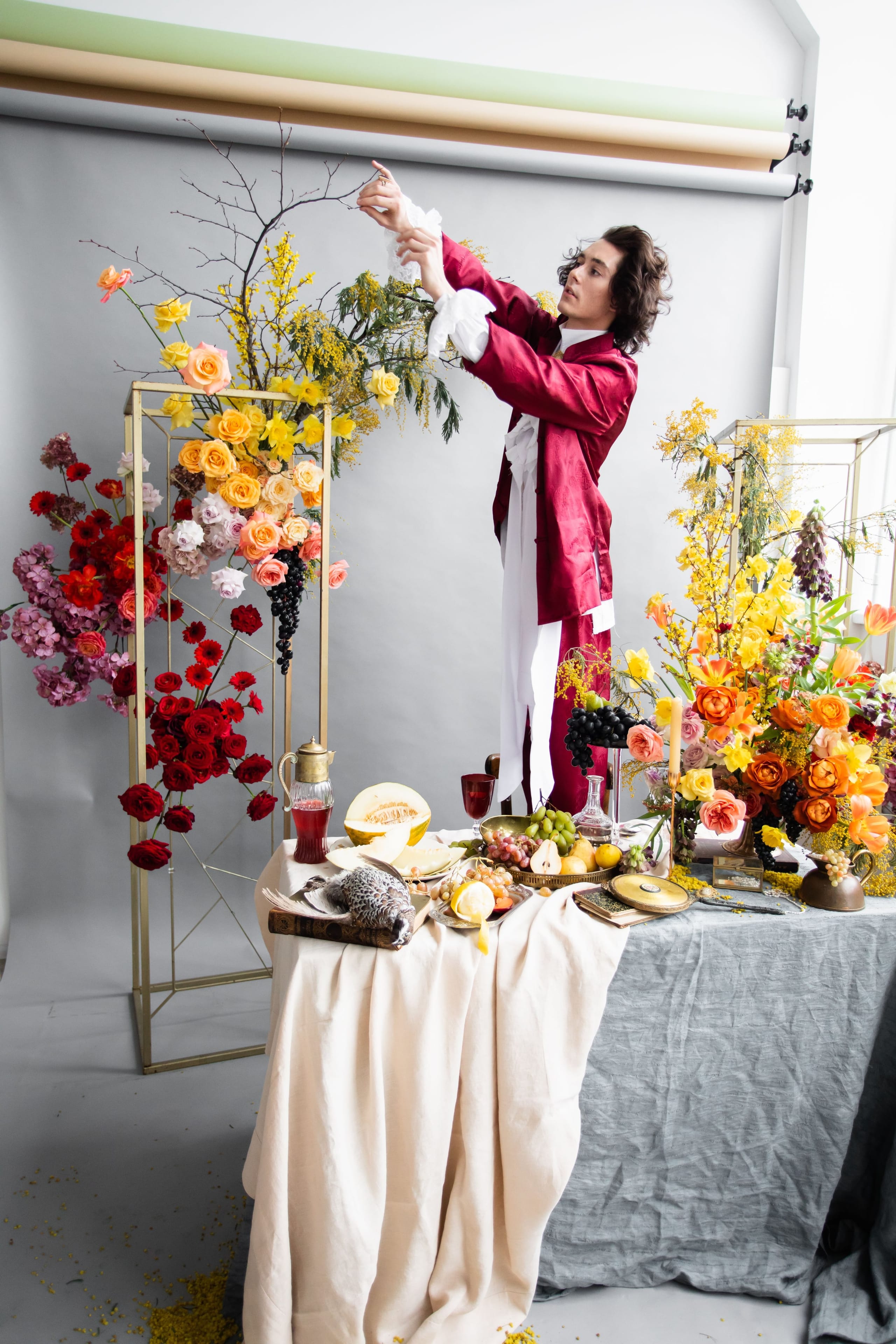 A man in a crimson satin coat with white lace cuffs reaches up to adjust a sprig of branches in a styled wedding table setting at a workshop in Russia. Around him, tall gold frames hold cascades of garden roses in red, peach and apricot, mauve hydrangea, dark grapes, and yellow forsythia and mimosa, with bright tulips and daffodils on the right. A melon, green grapes, a glass decanter and candles sit on the table below in warm, golden-toned light.