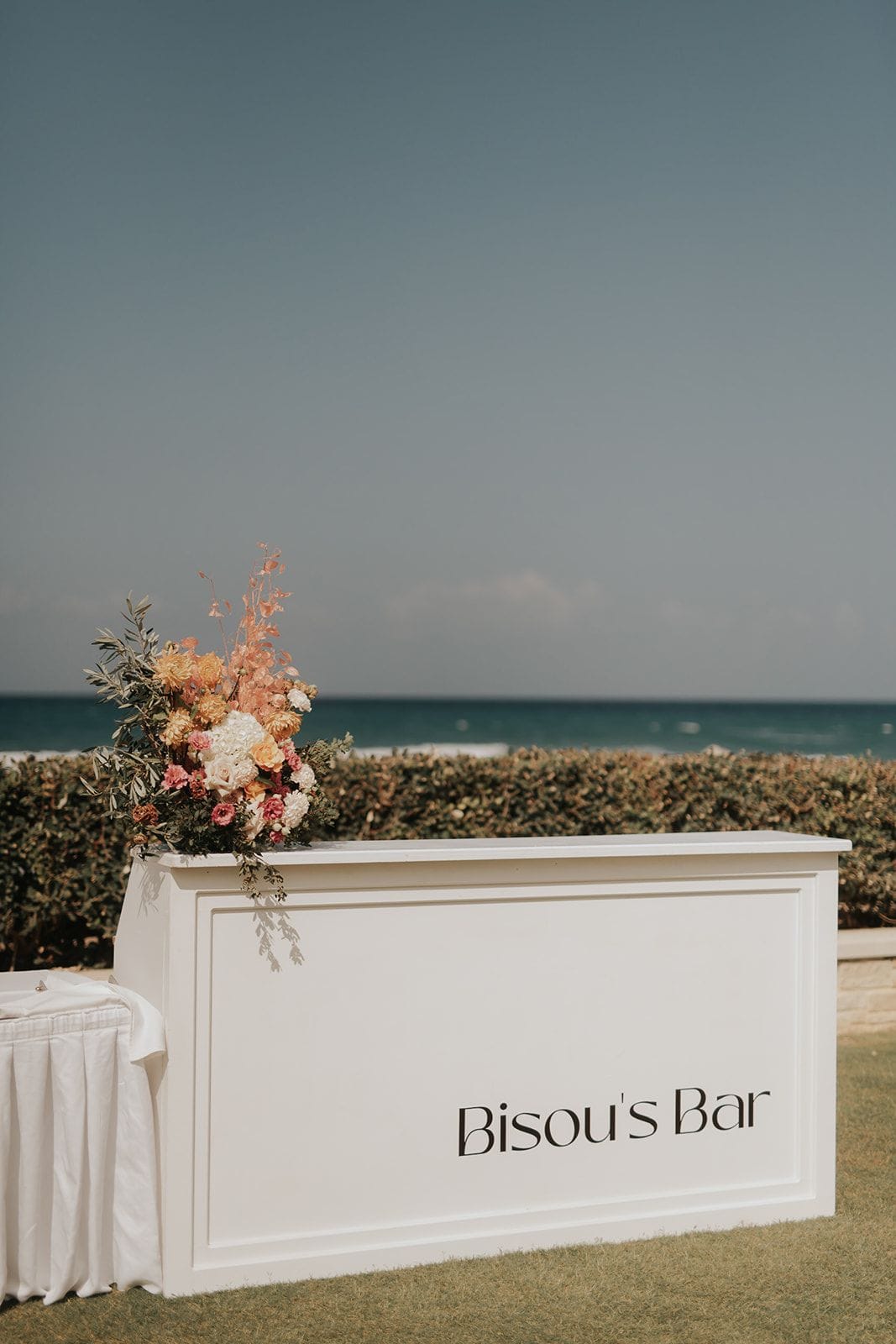 A white wooden bar with a panelled front, lettered "Bisou's Bar" in black, stands on a lawn beside a clipped hedge and a turquoise sea under a grey sky. On its top sits a tall arrangement of apricot and peach dahlias, white hydrangea, blush roses and pink carnations, with olive branches and trailing eucalyptus spilling over the edge. This wedding flower display in Greece has a soft, sun-warmed palette that echoes the seaside setting.