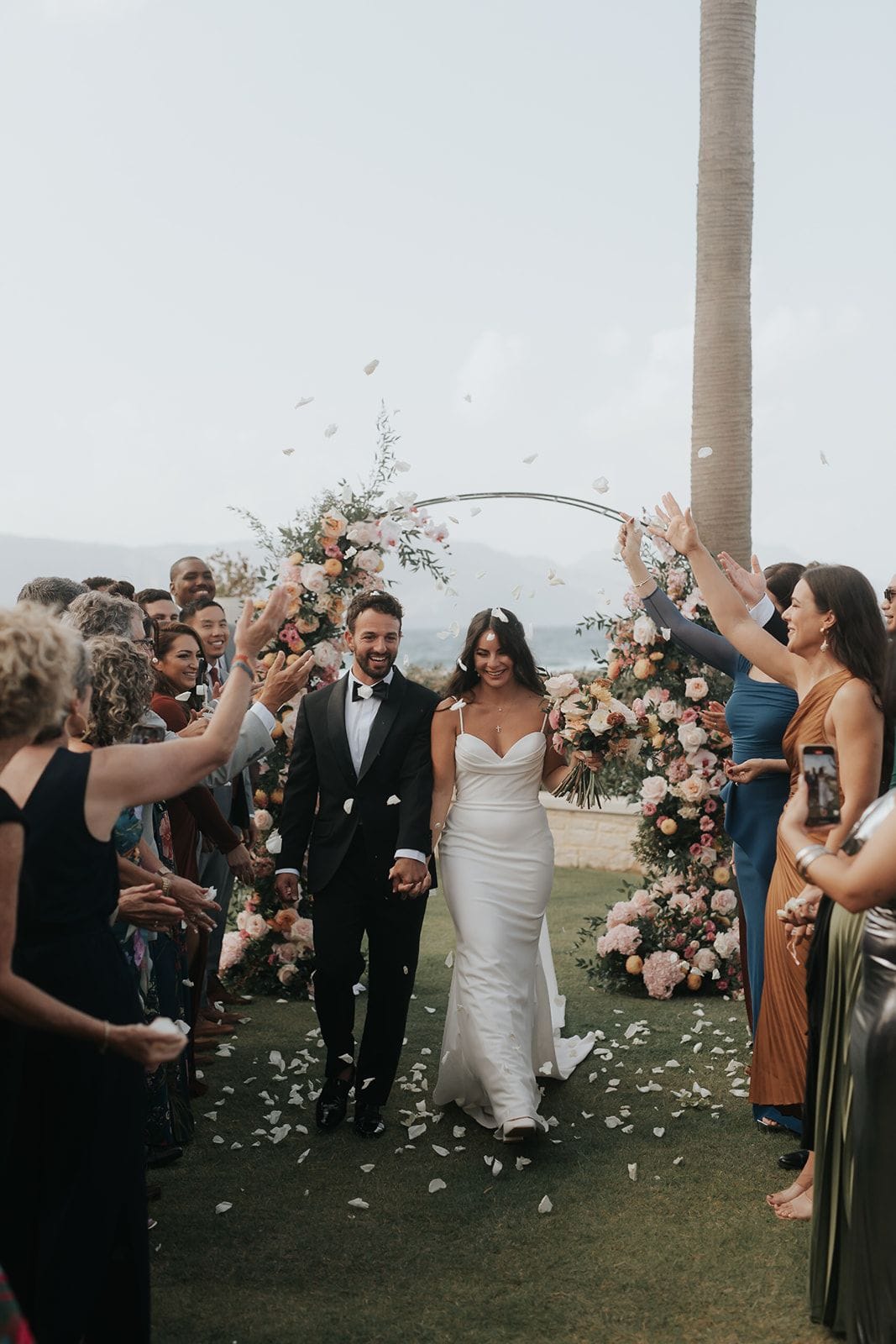 A bride and groom walk hand in hand down a grassy aisle while guests line both sides, raising their arms and tossing white petals into the air. Behind them stands a tall circular arch wrapped in pink and peach garden roses, blush peonies and greenery, with a hazy sea and mountains beyond, a wedding setting in Greece.