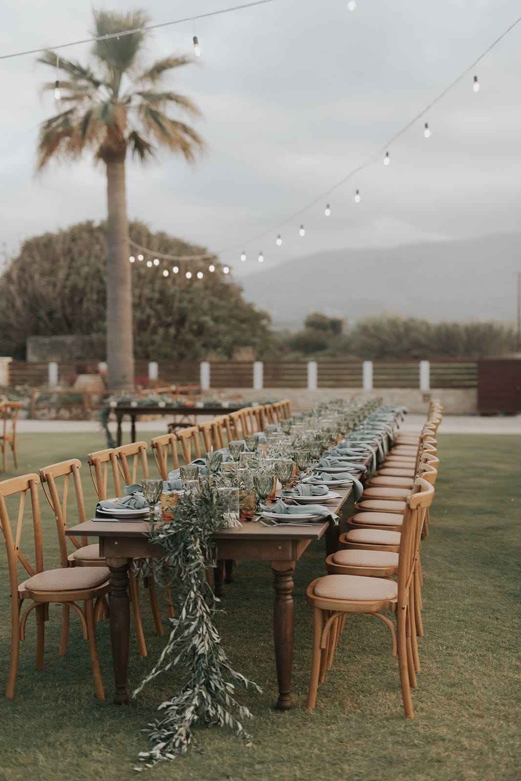 A long wedding dining table set outdoors on a lawn in Greece, with rows of wooden cross-back chairs along both sides. Dusty blue napkins sit on white plates beside tall green-tinted glasses, and a runner of olive branches and eucalyptus stretches down the centre. Strings of warm bulbs hang overhead beneath a soft grey sky, with a tall palm and a misty mountain behind.