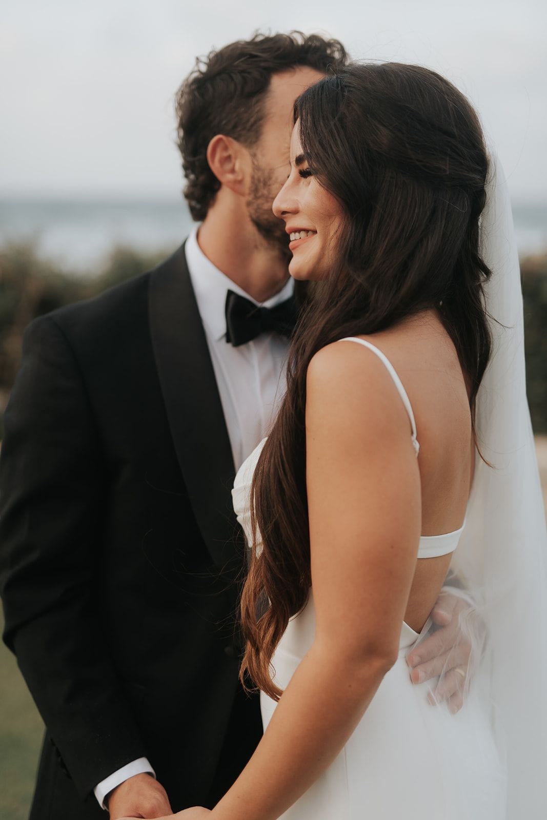 A close, soft-focus portrait of a bride and groom standing side by side, the groom in a black tuxedo with a black bow tie and white shirt, his dark curly hair and stubble close to the bride's head as he leans in. The bride, with long dark wavy hair and a sheer veil falling behind her, smiles warmly with her face turned away from the camera toward the sea. Behind them the sky is pale and hazy with blurred greenery at the edges, a wedding portrait taken in Greece.