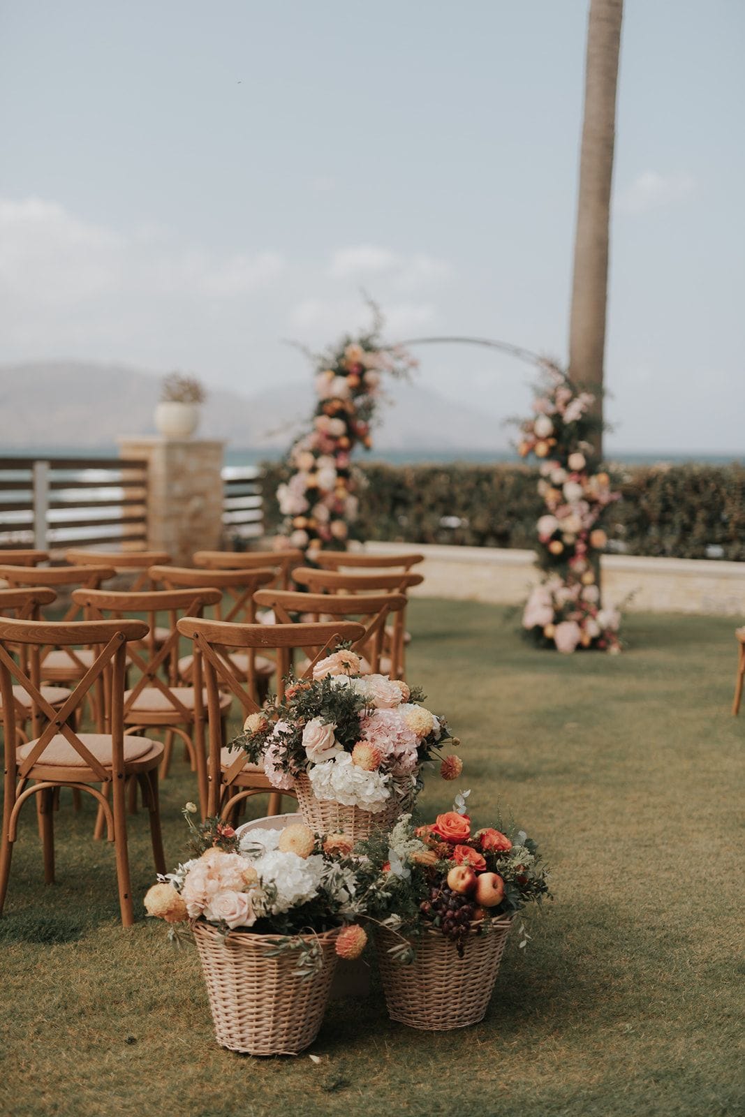 Three woven wicker baskets of flowers sit on the lawn at the end of a row of wooden cross-back chairs, with blush garden roses, white hydrangeas, peach dahlias and olive sprigs in the front pair, and orange roses, apples and grapes in the one beside them. Behind them, tall floral columns in blush and peach tones frame an aisle that runs toward the sea, with a stone wall and hedge beyond. This wedding flower display is in Greece, set outdoors in soft, hazy daylight.